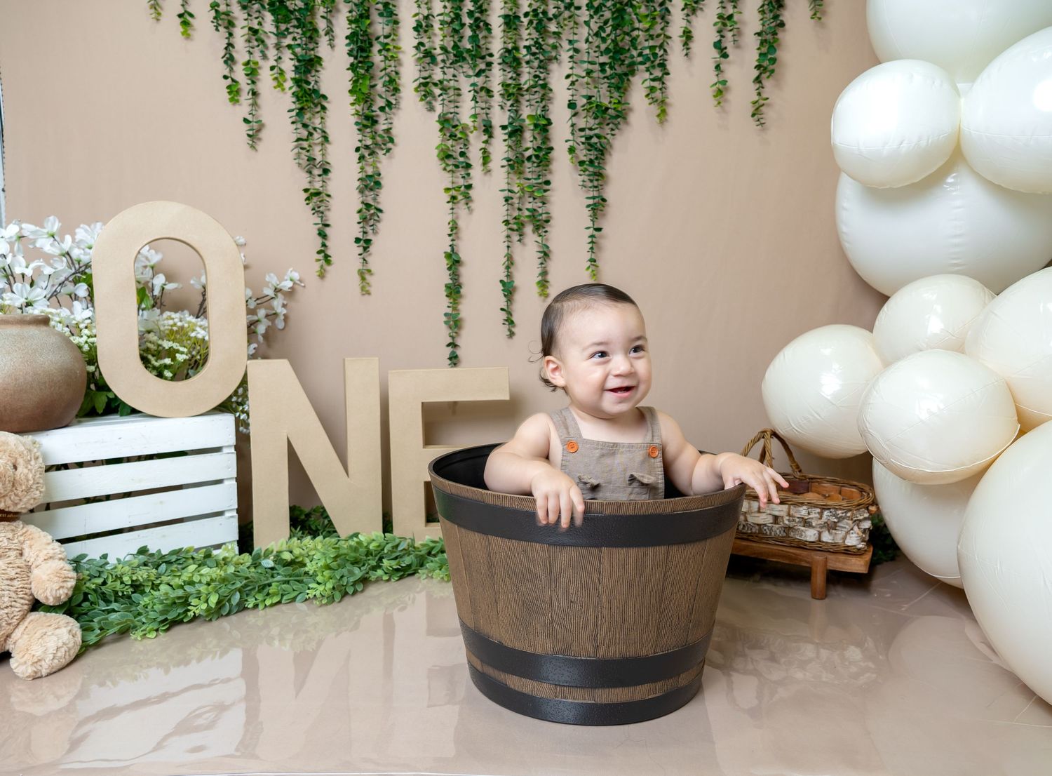 Baby sitting in a wooden barrel tub at a first birthday party with ONE letters, white balloons, and green ivy backdrop.