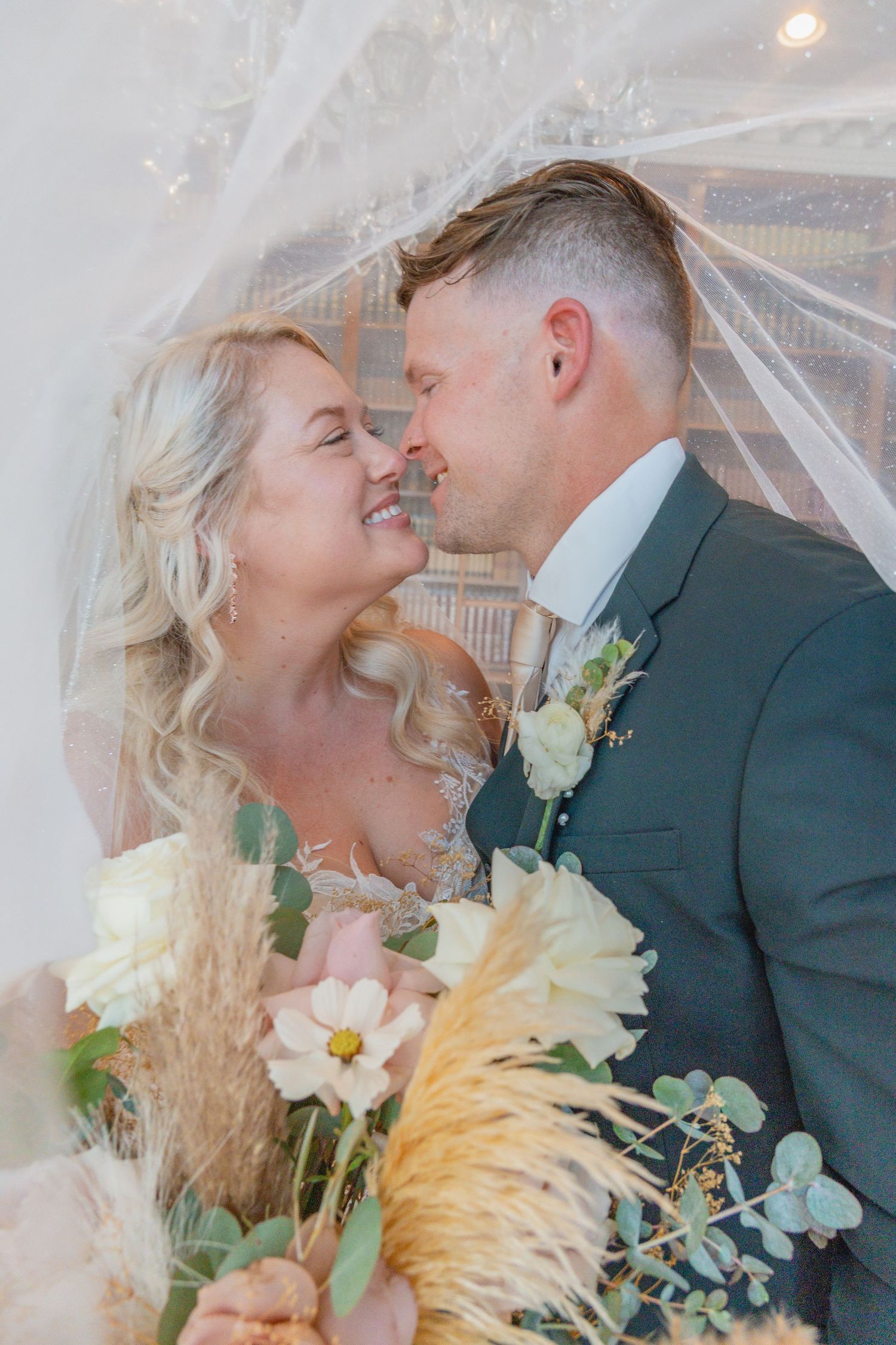 Bride and groom share a romantic moment under a veil, with a bouquet of white and cream flowers.