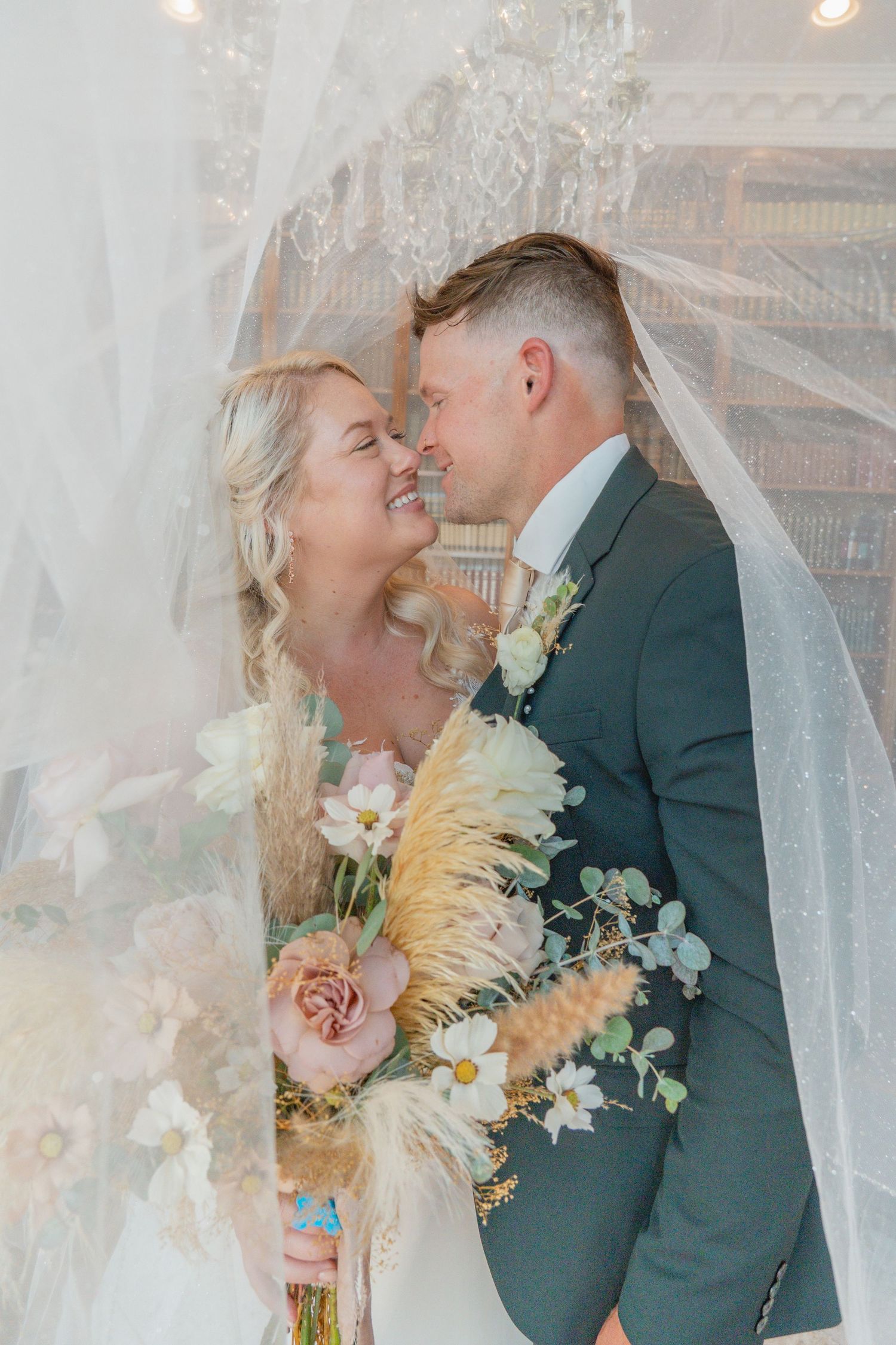 Smiling bride and groom share a romantic moment under a veil, holding a boho bouquet in a library.