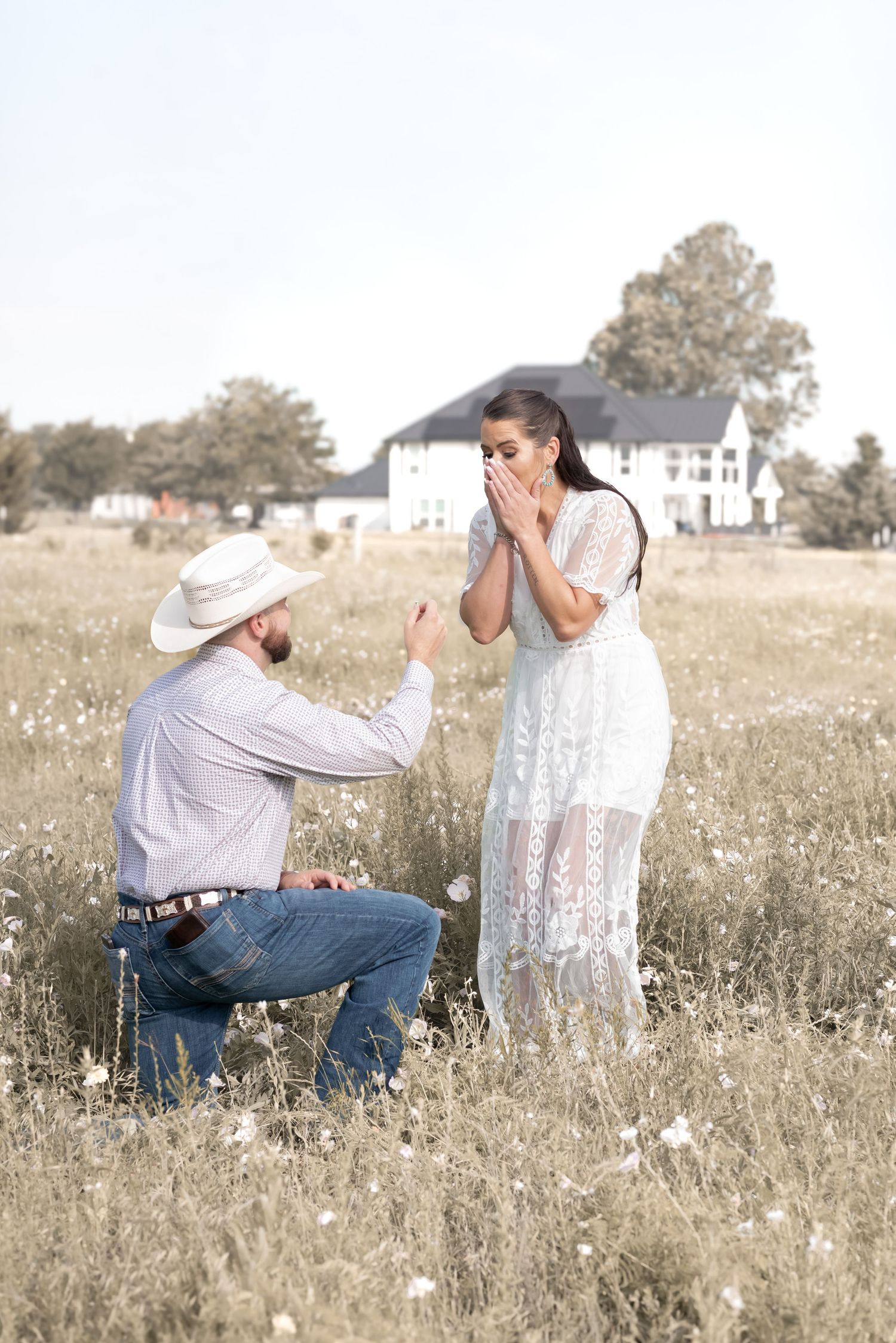 Man in cowboy hat proposes to surprised woman in white lace dress in a wildflower field.