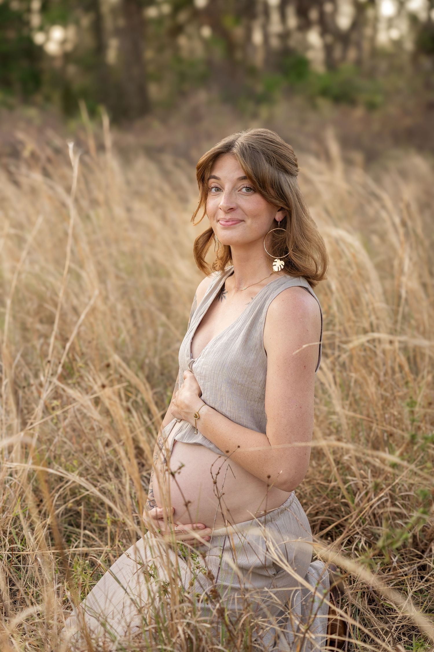 Pregnant woman in gray linen dress holding baby bump while standing in tall golden grass outdoors.