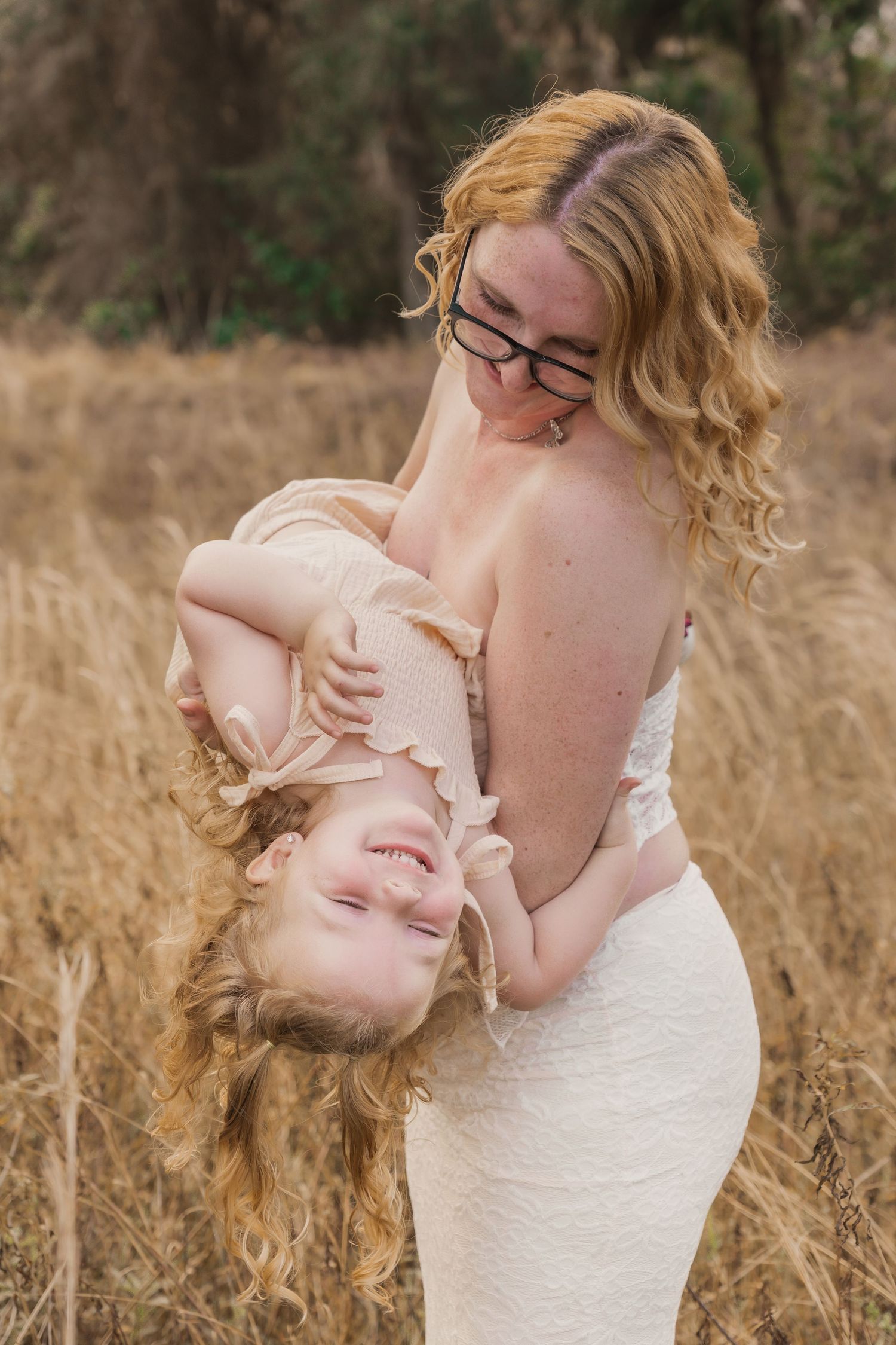 A mother playfully holds her laughing toddler upside down in a golden field during a outdoor photo session.