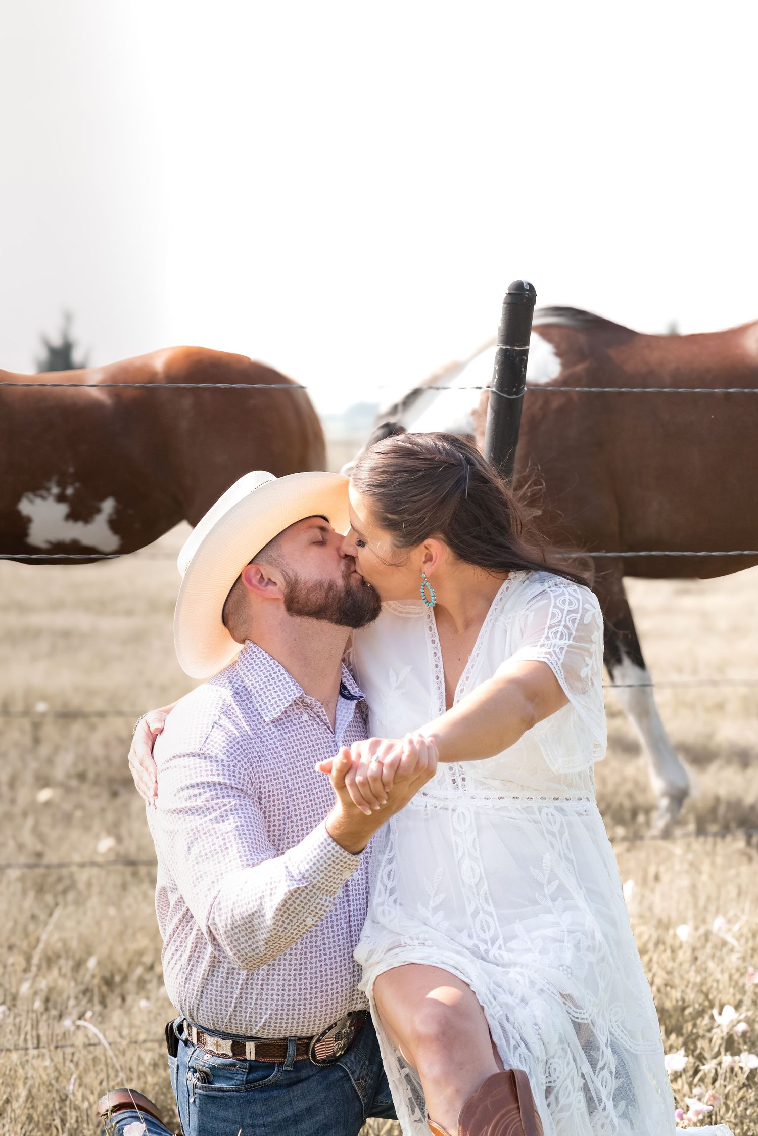 Couple sharing a romantic kiss on a ranch with horses behind them, man wearing cowboy hat and woman in white lace dress.