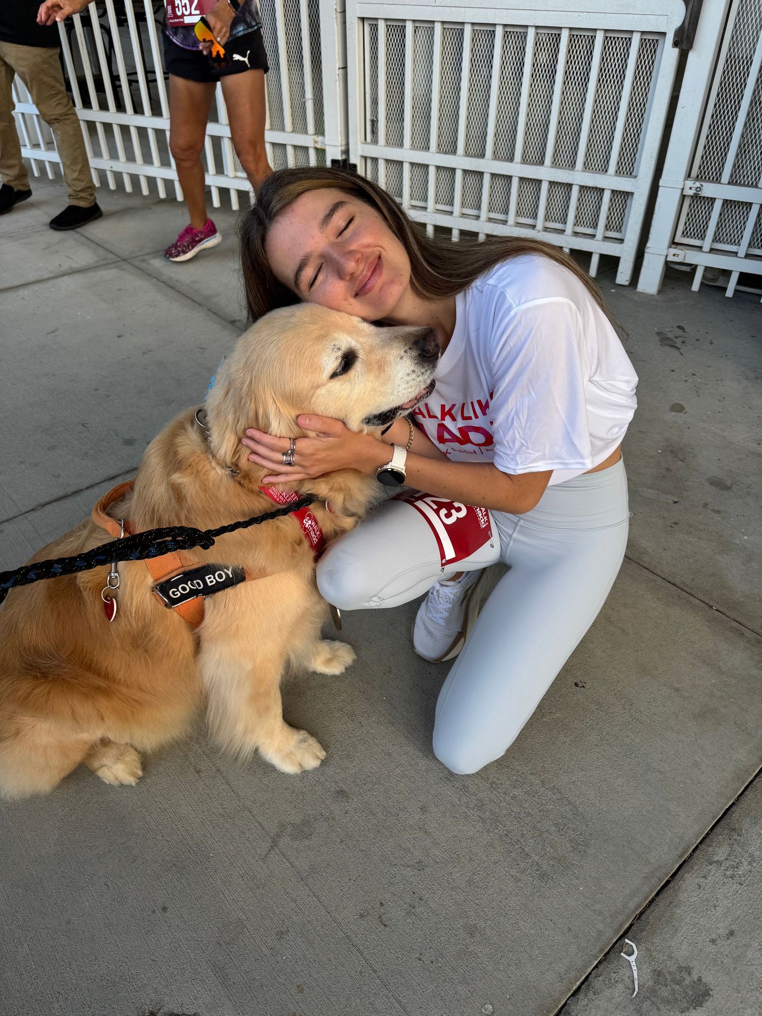 A smiling girl hugs a golden retriever service dog wearing a red vest outdoors.