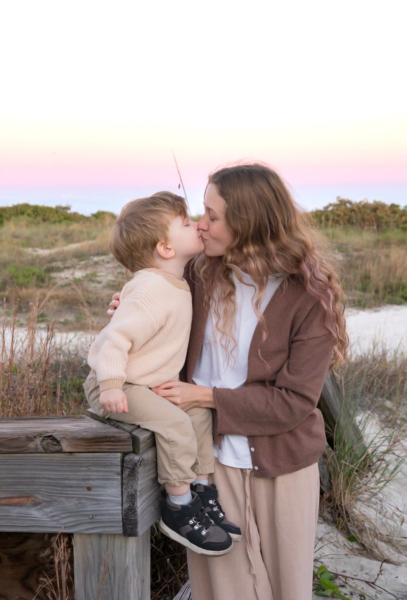 Tender moment of mother and toddler son kissing at sunset beach with pastel sky backdrop.