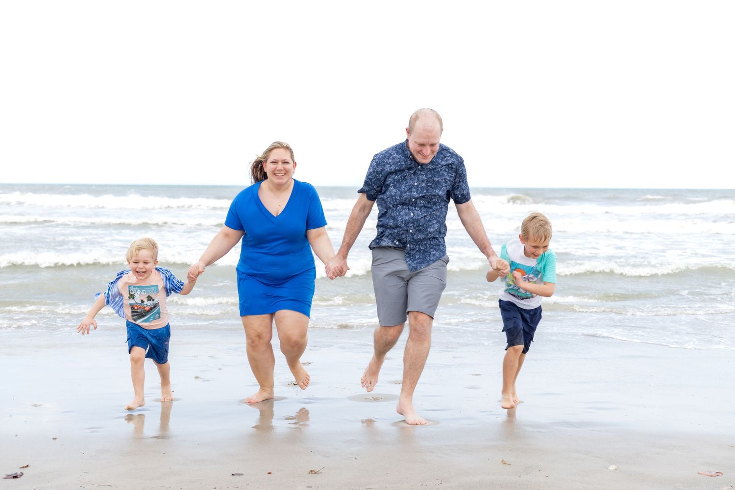 Family of four running barefoot on the beach, parents holding hands with two young boys near ocean waves.