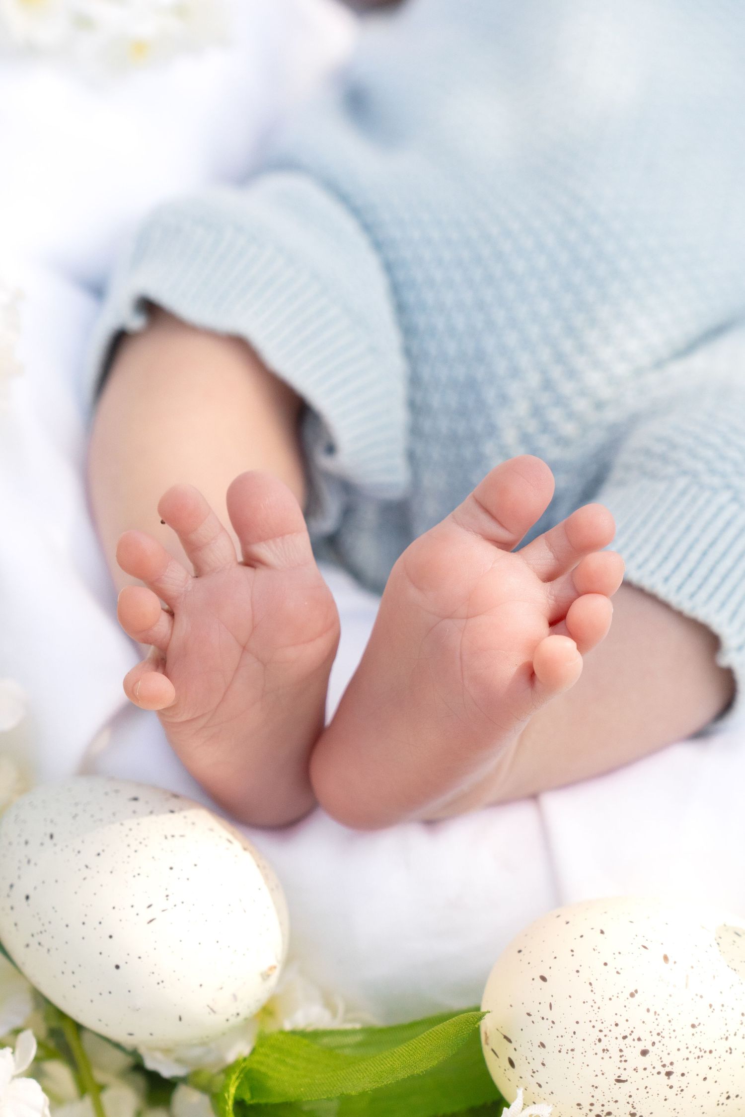 Tiny newborn baby feet in blue knit outfit surrounded by white speckled Easter eggs and spring flowers.