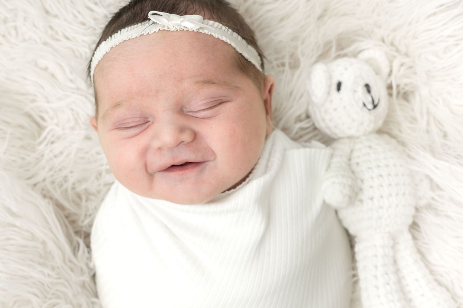 Smiling newborn baby girl wrapped in white swaddle with headband, lying next to a crocheted teddy bear on fluffy rug.