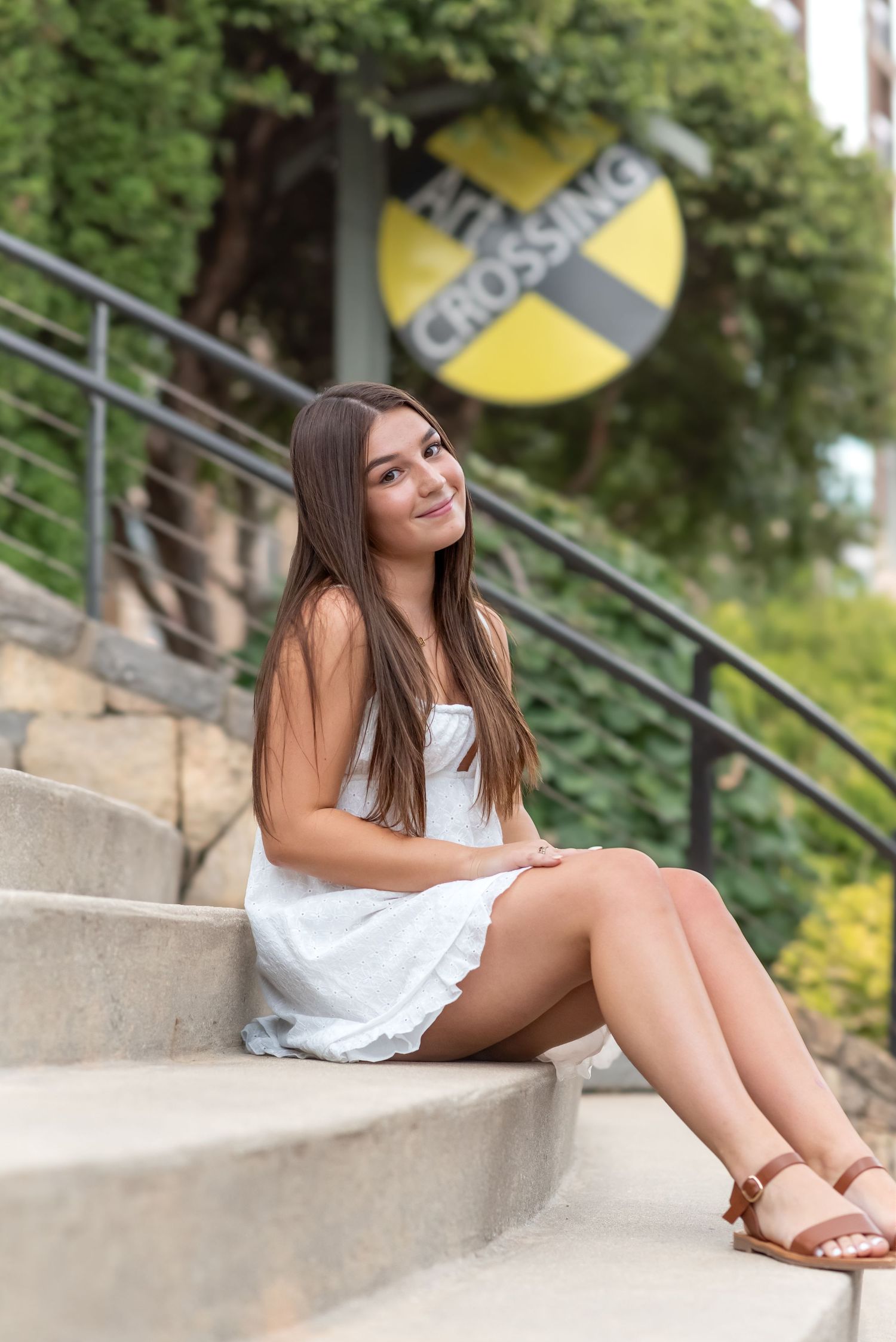 Young woman in white dress sitting on outdoor steps near a railroad crossing sign, smiling at camera.