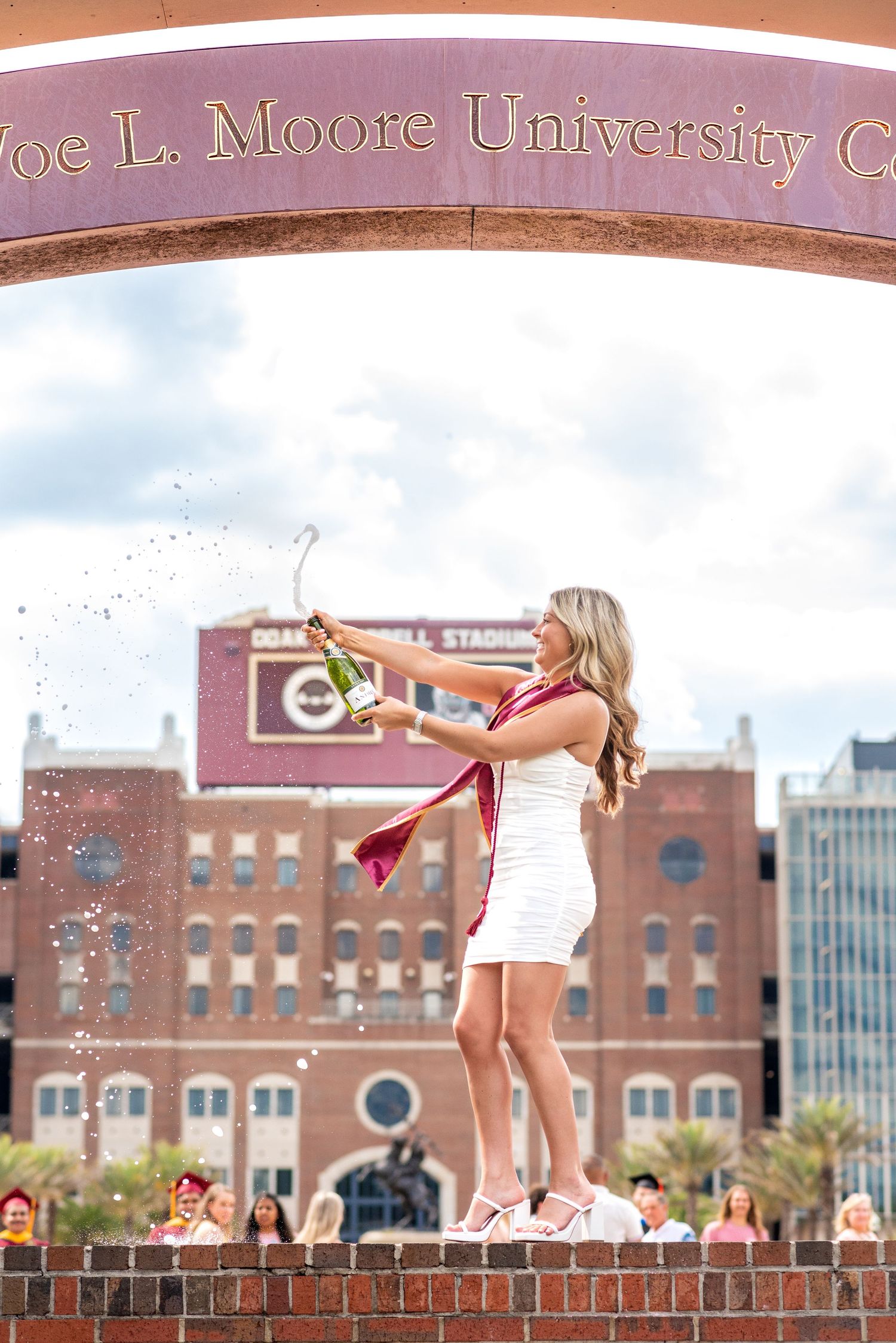 A blonde woman in a white dress pops champagne under the Joe L. Moore University arch at Florida State University.