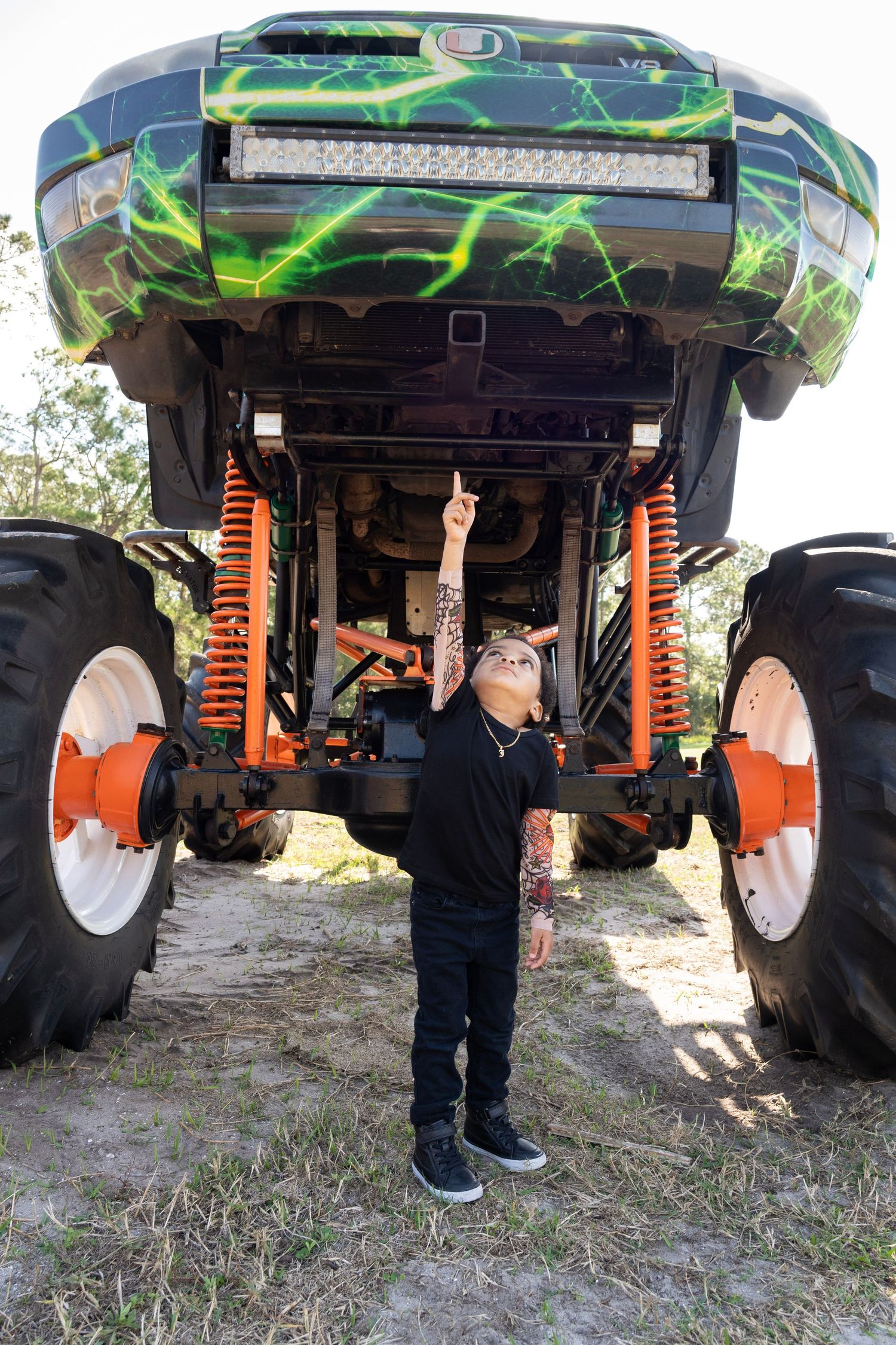 Young boy pointing up at the undercarriage of a large green monster truck with orange suspension and oversized tires.
