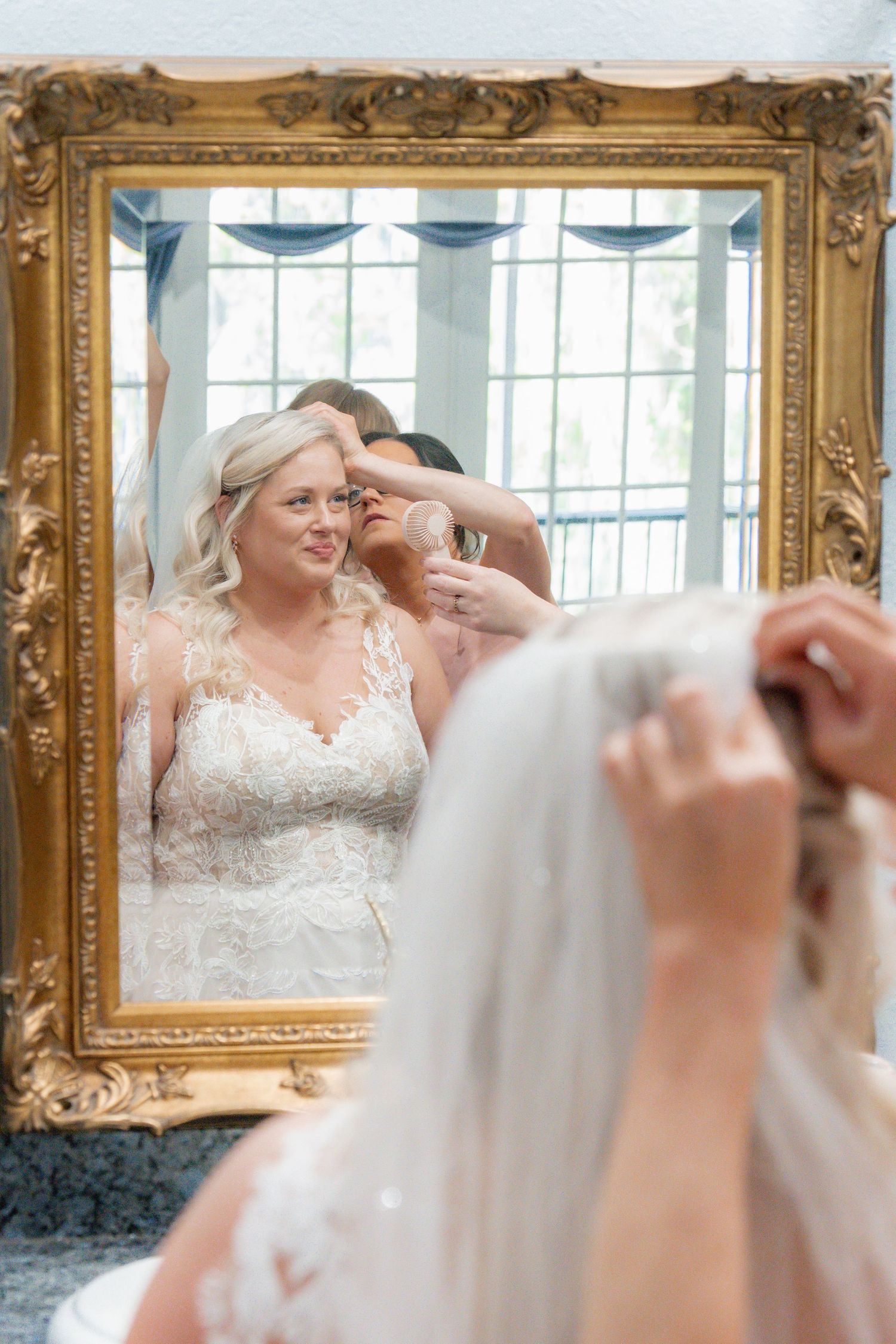 Bride in lace wedding gown smiles in ornate gold mirror as bridesmaid adjusts her veil.