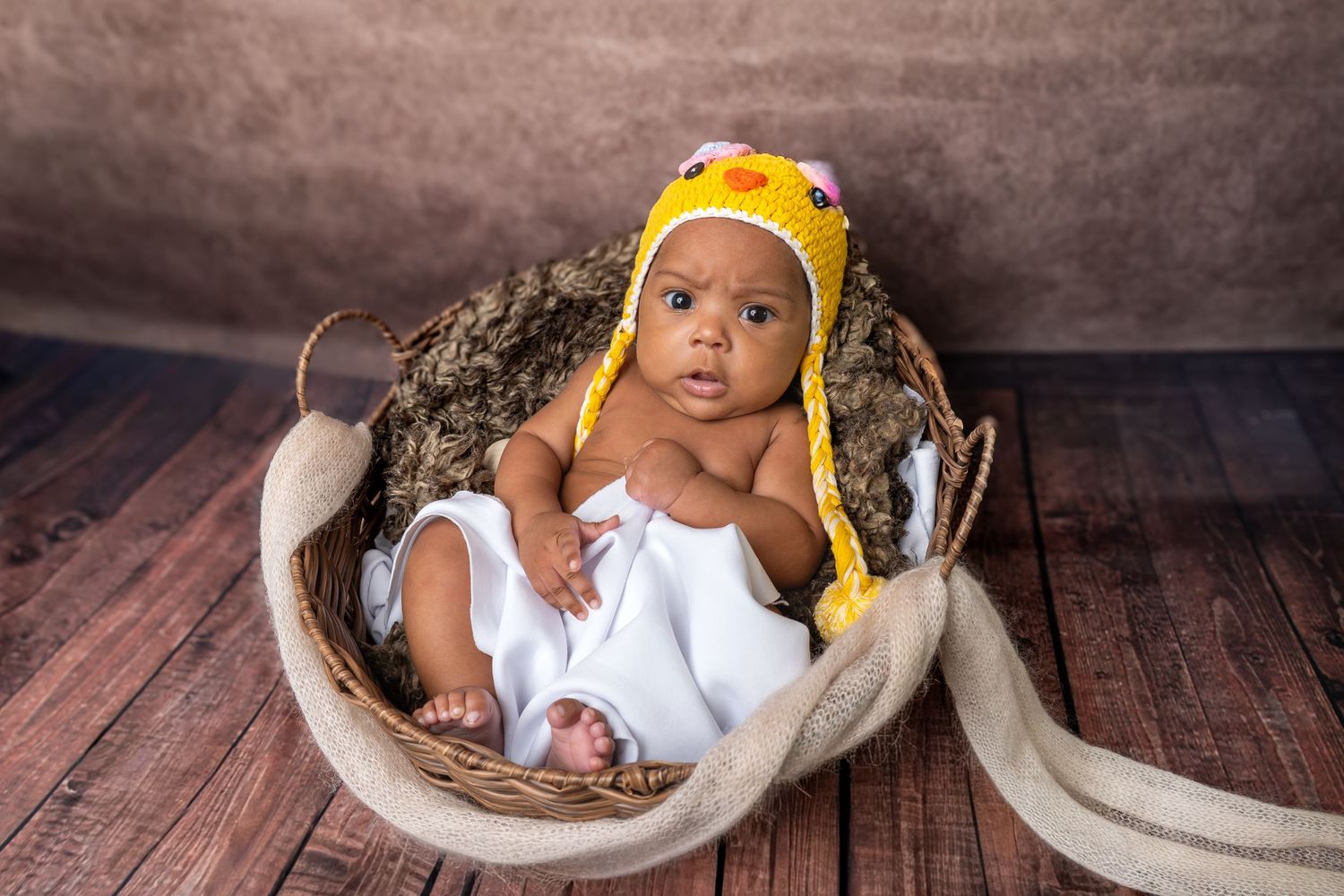 Newborn baby wearing a yellow crocheted hat sitting upright in a woven basket on a wooden floor.