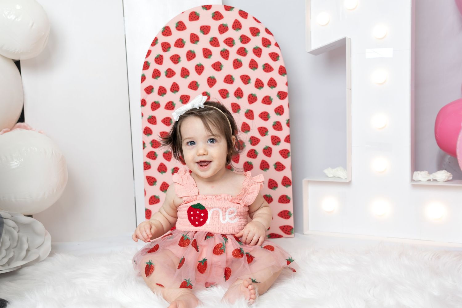 Baby girl in strawberry dress sits smiling before a strawberry arch backdrop with balloon decorations.