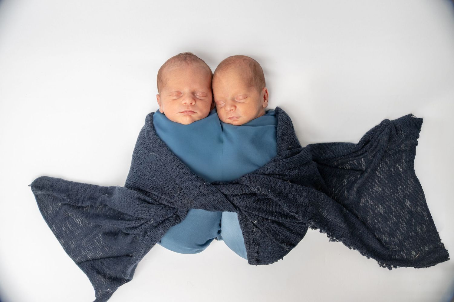Two sleeping newborn twins swaddled together in blue and dark navy wraps on a white background.