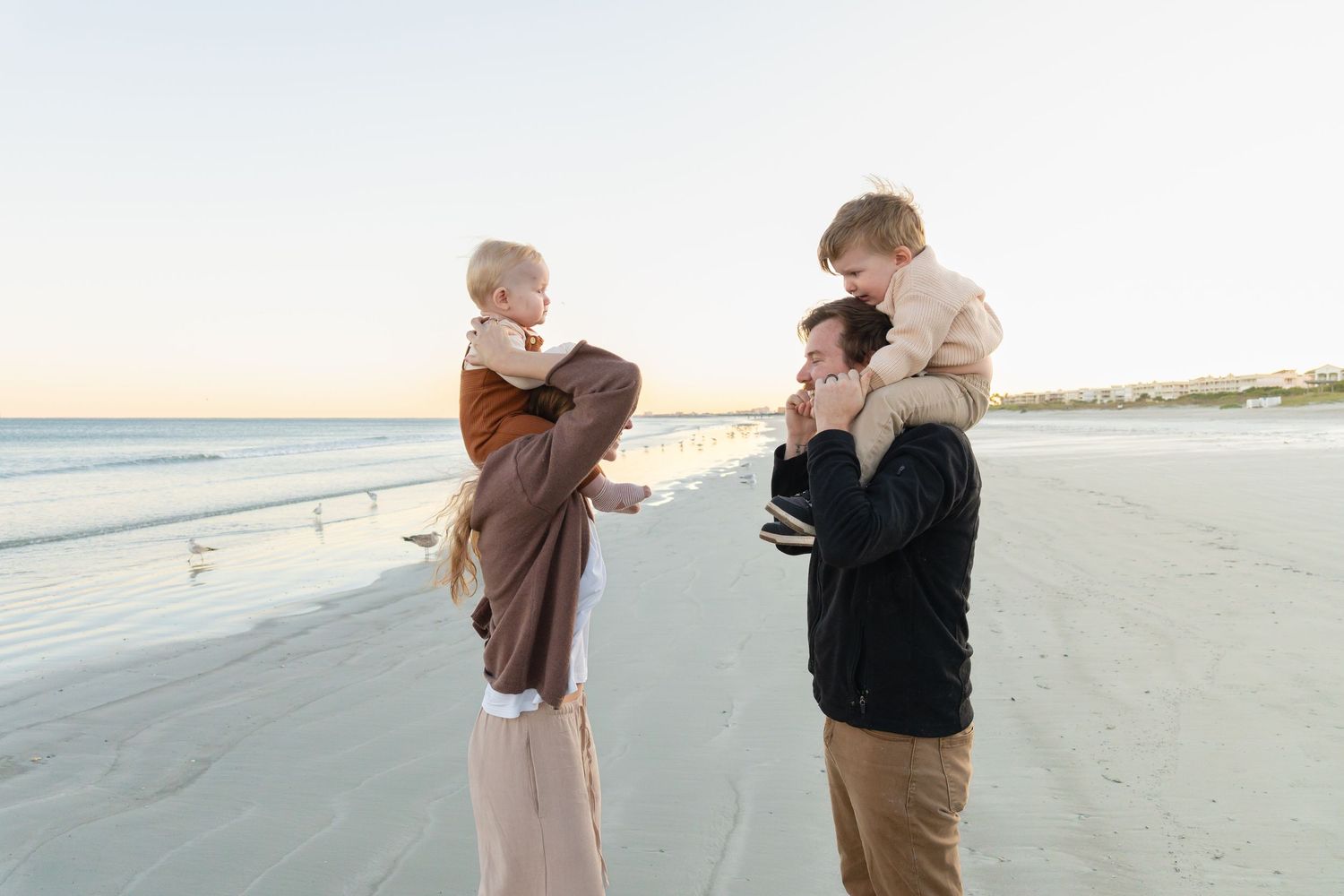 Family of four enjoying a beach day, with parents each holding a young child on a sandy shore at sunset.