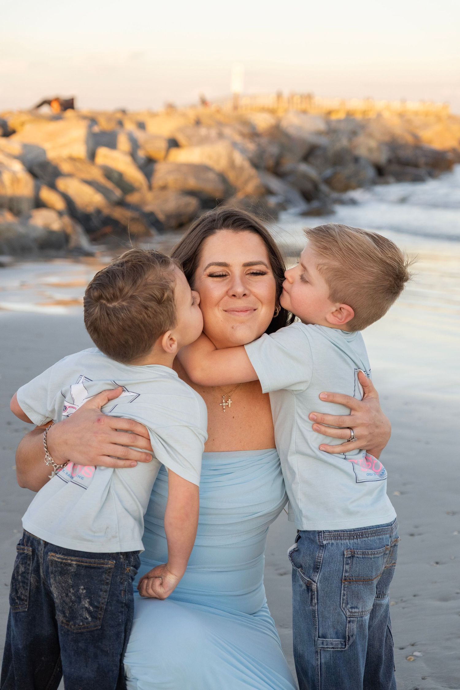 A mother smiles as her two young sons kiss her cheeks on a sandy beach at sunset.