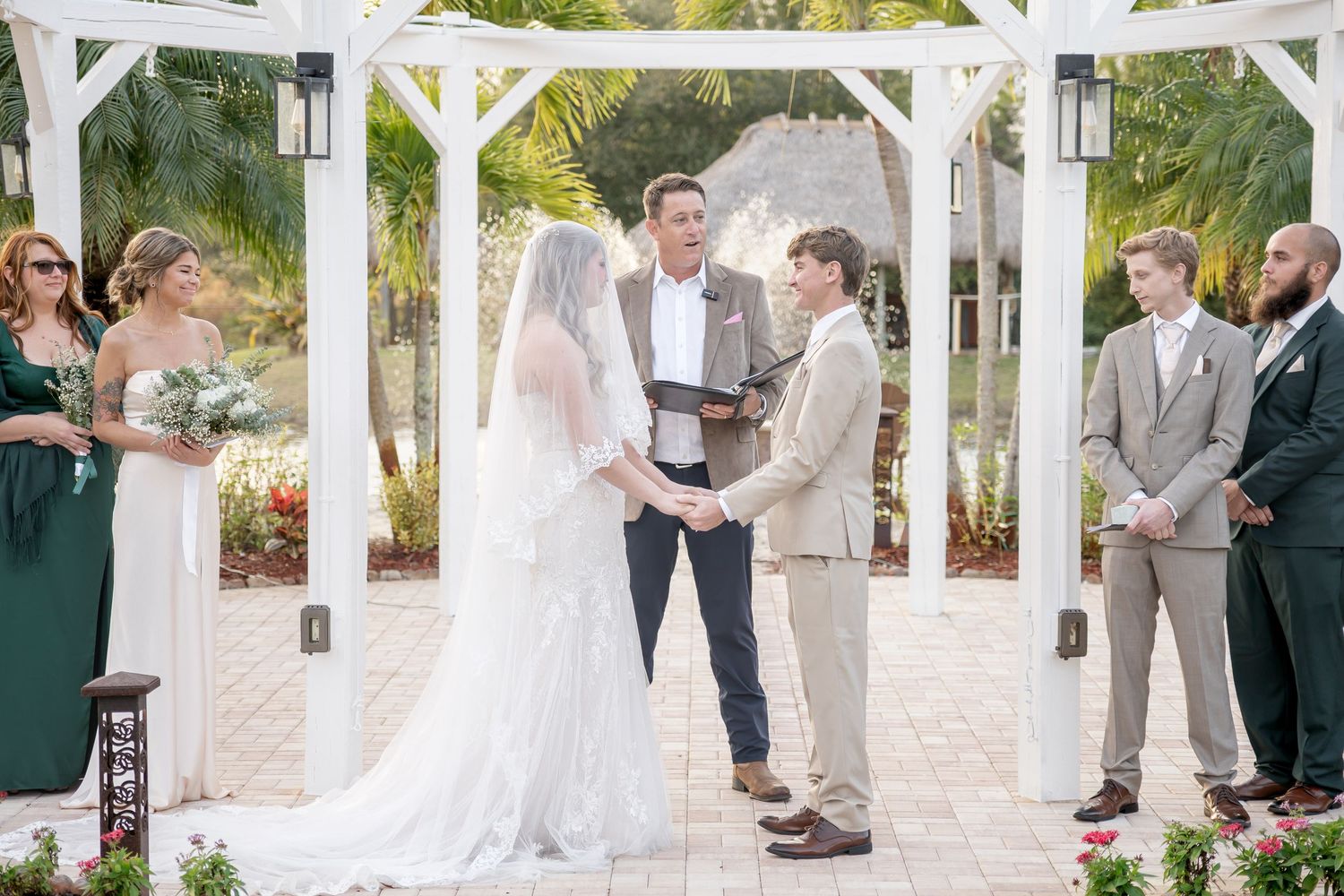 Bride and groom exchanging vows under a white gazebo surrounded by wedding guests outdoors.