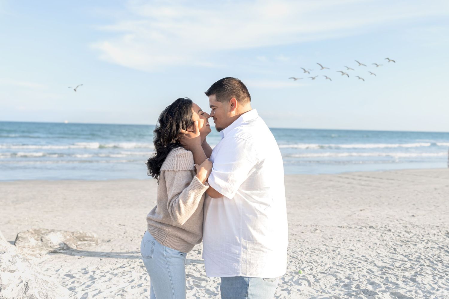 Romantic couple embracing on white sand beach with ocean waves and birds flying in background.