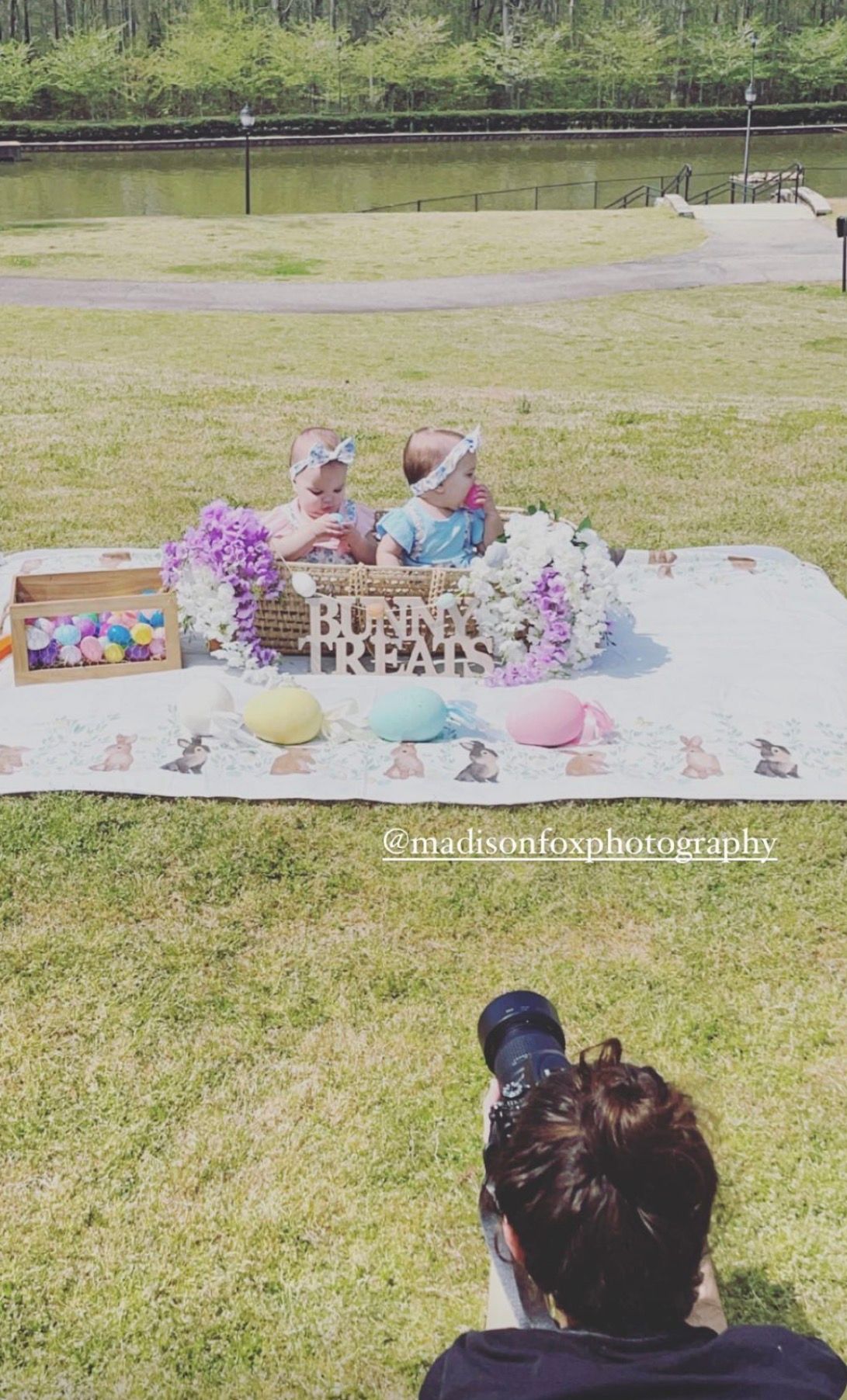 Photographer captures two babies in Easter-themed outdoor photoshoot with bunny treats sign and colorful eggs.