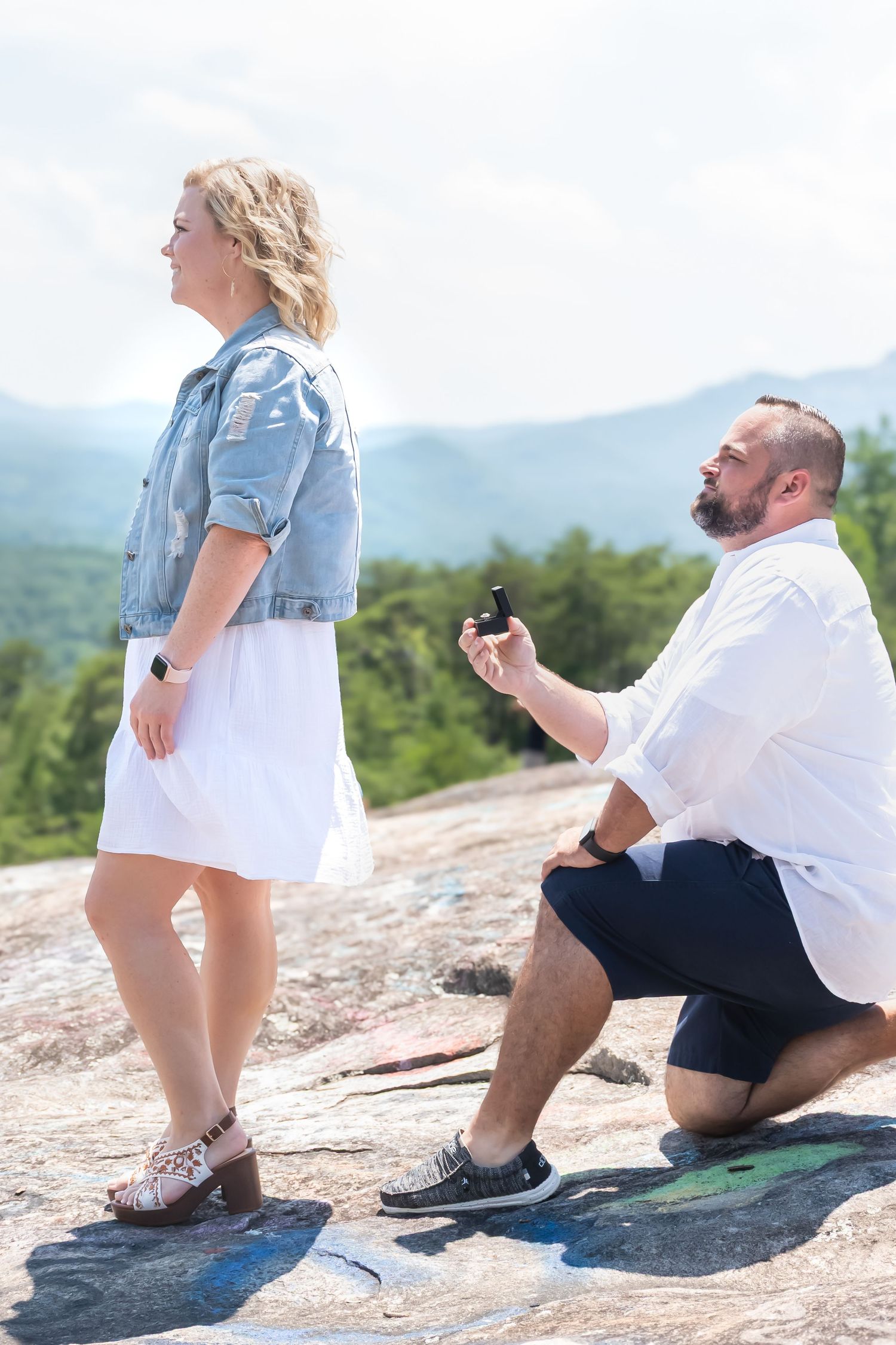 Man kneeling proposing to woman on granite rock with mountain landscape in background.