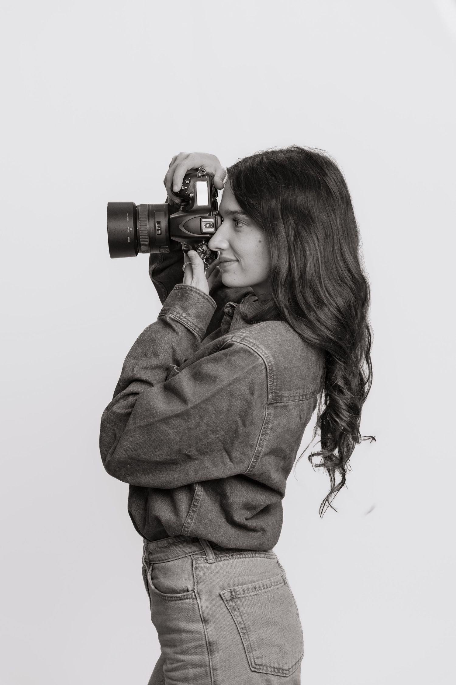 Black and white photo of young woman in denim jacket photographing with DSLR camera sideways.