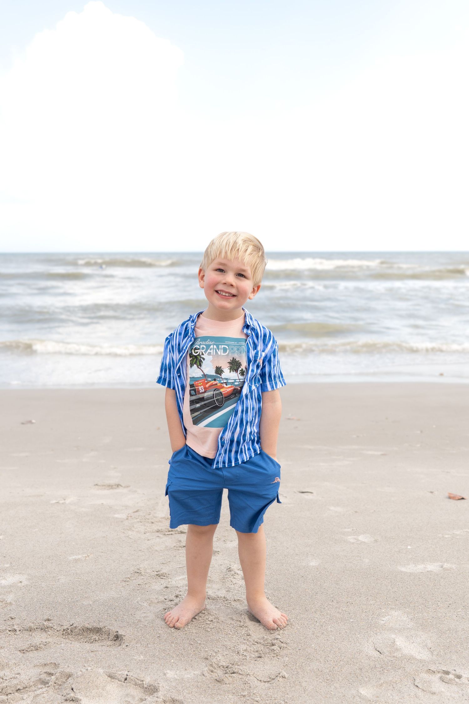 Smiling blonde boy in blue shorts and striped shirt standing barefoot on sandy beach with ocean waves in background.
