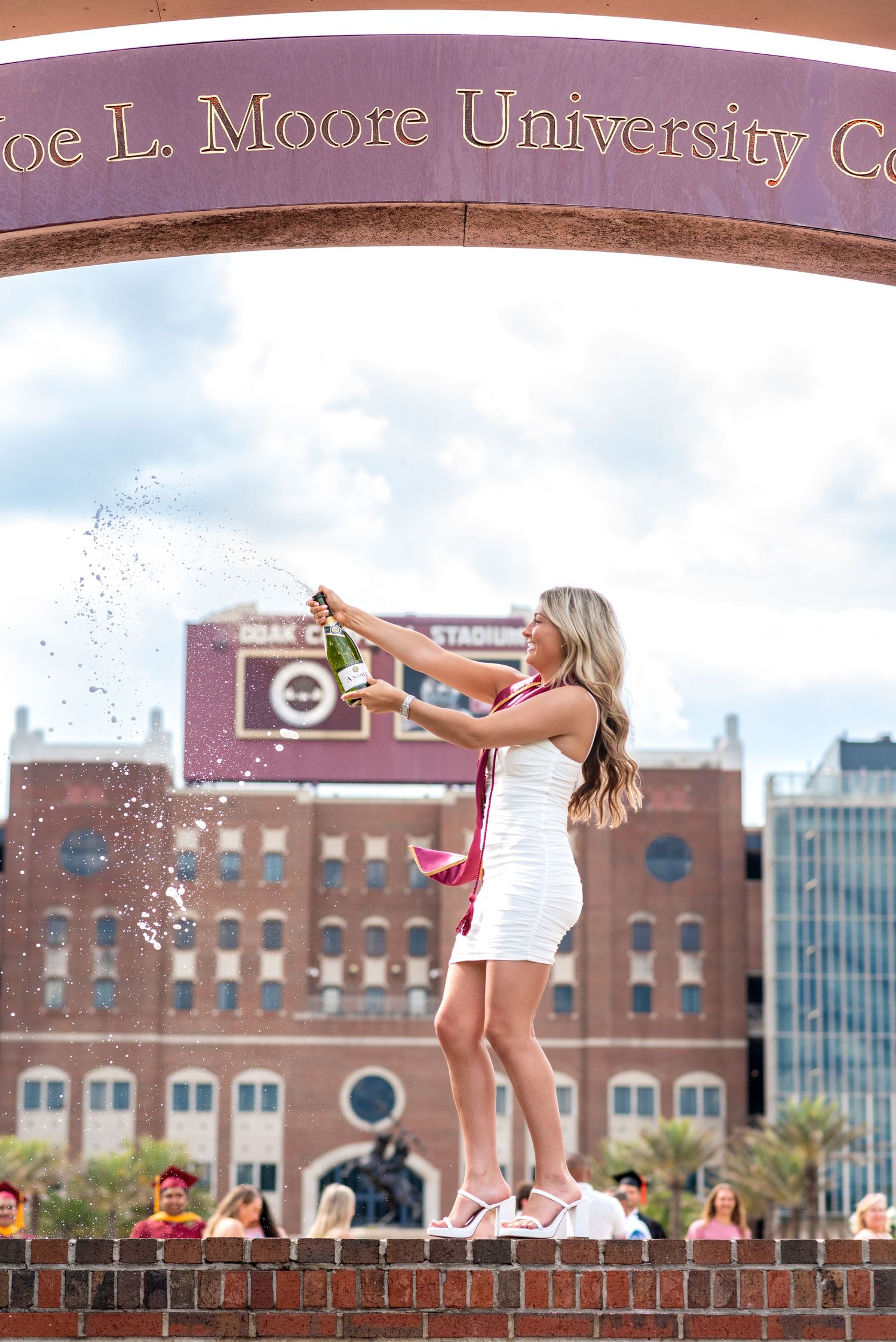 Graduate in white dress pops champagne under Joe L. Moore University arch with Doak Campbell Stadium behind her.