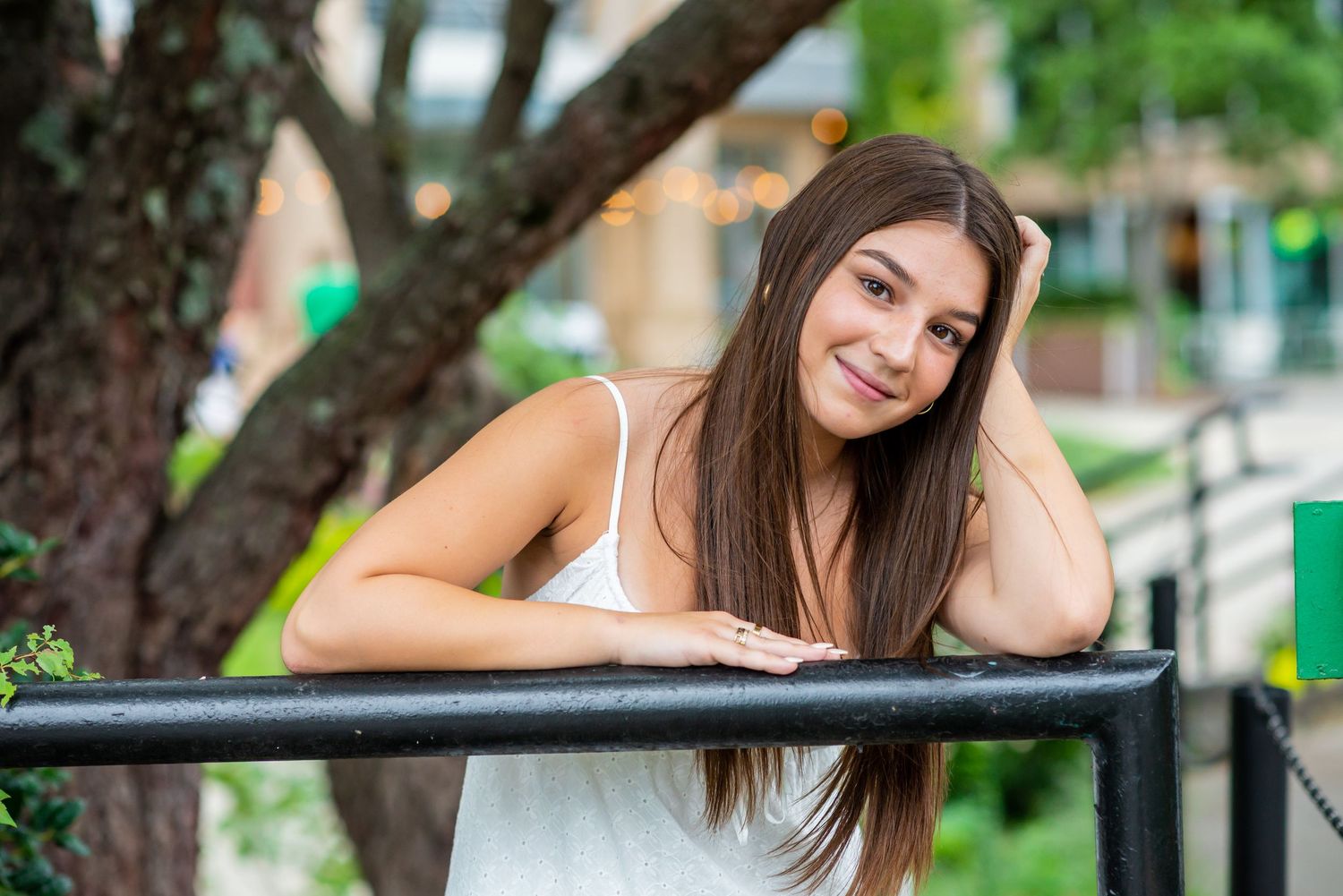 Smiling young woman with long brown hair leaning on a black railing outdoors in a white sleeveless top.