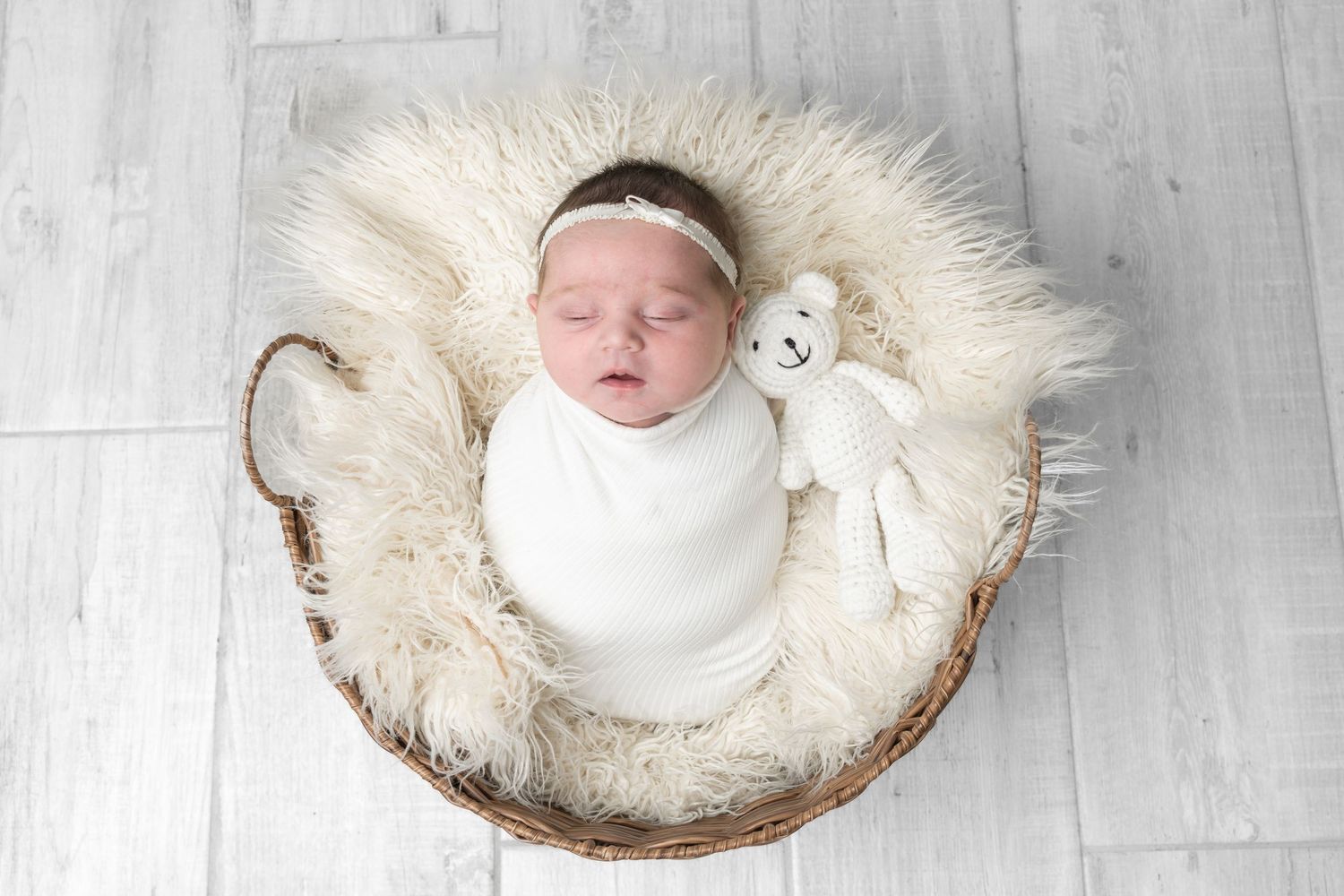 Newborn baby girl swaddled in white, sleeping peacefully in a wicker basket with fluffy fur lining and teddy bear.