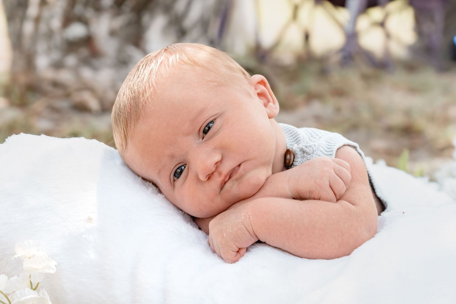 Newborn baby resting on a white blanket outdoors, with hands tucked under chin, looking at the camera.