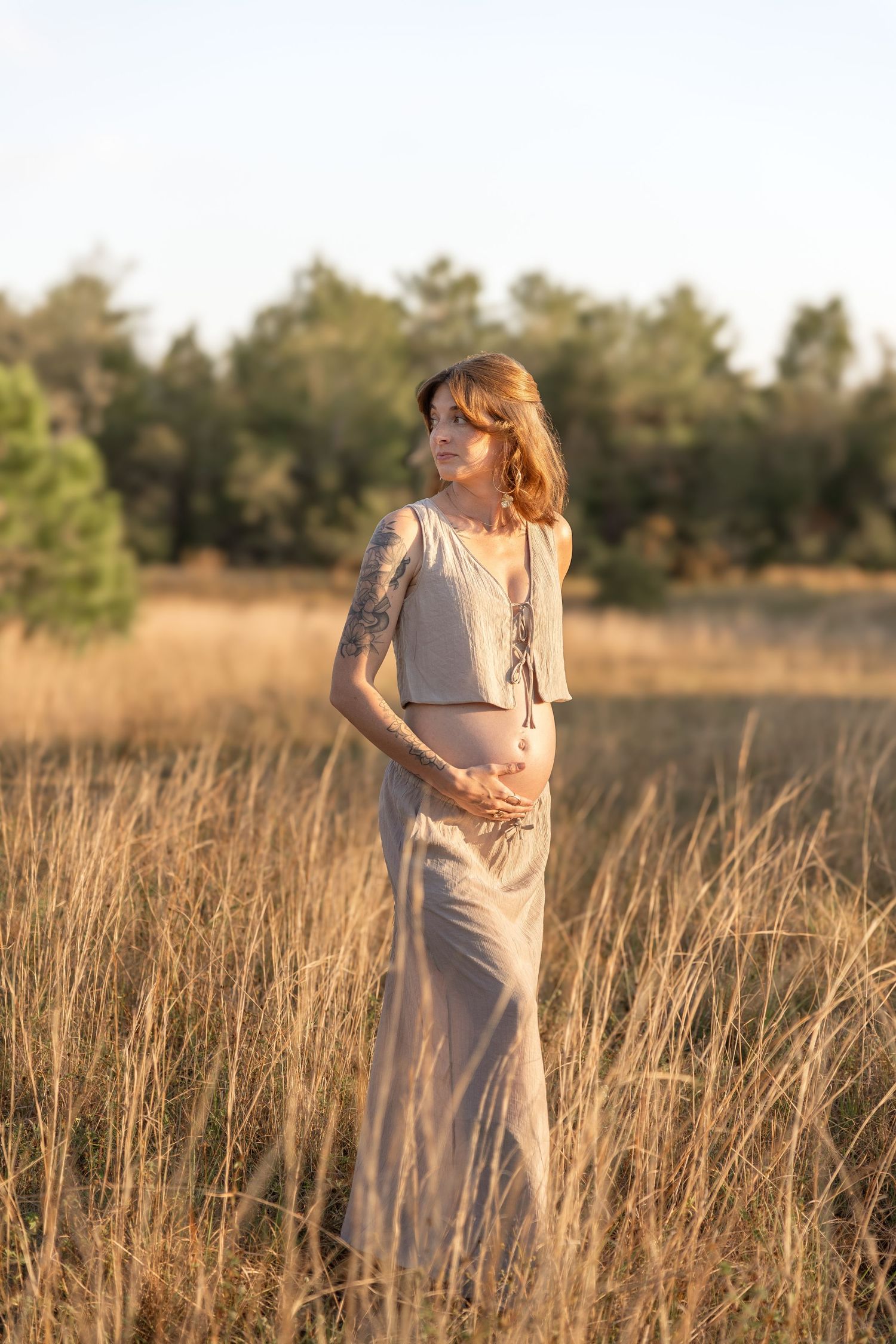 Pregnant woman in beige outfit standing in golden field at sunset, showing baby bump with tattoos visible.