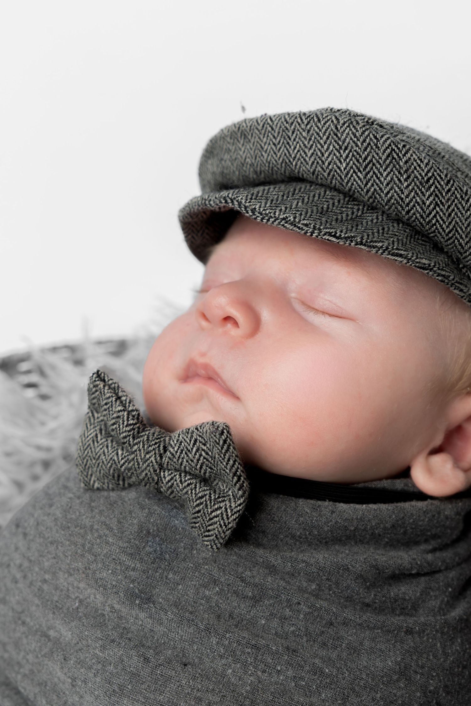 Sleeping newborn baby wearing a gray herringbone flat cap and matching bow tie, wrapped in a soft gray blanket.