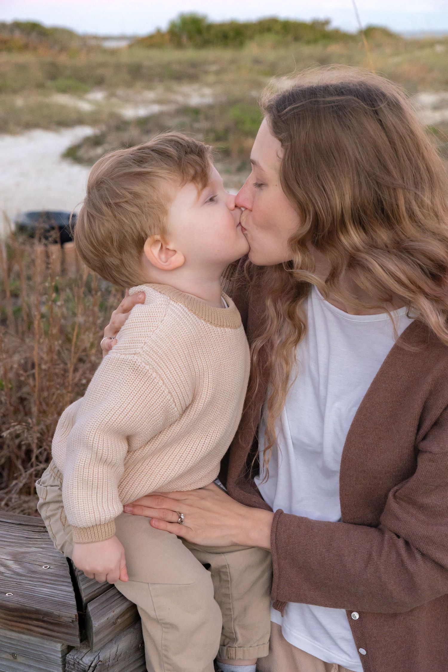 A mother kissing her toddler son on wooden steps near a sandy beach, both wearing neutral tones.