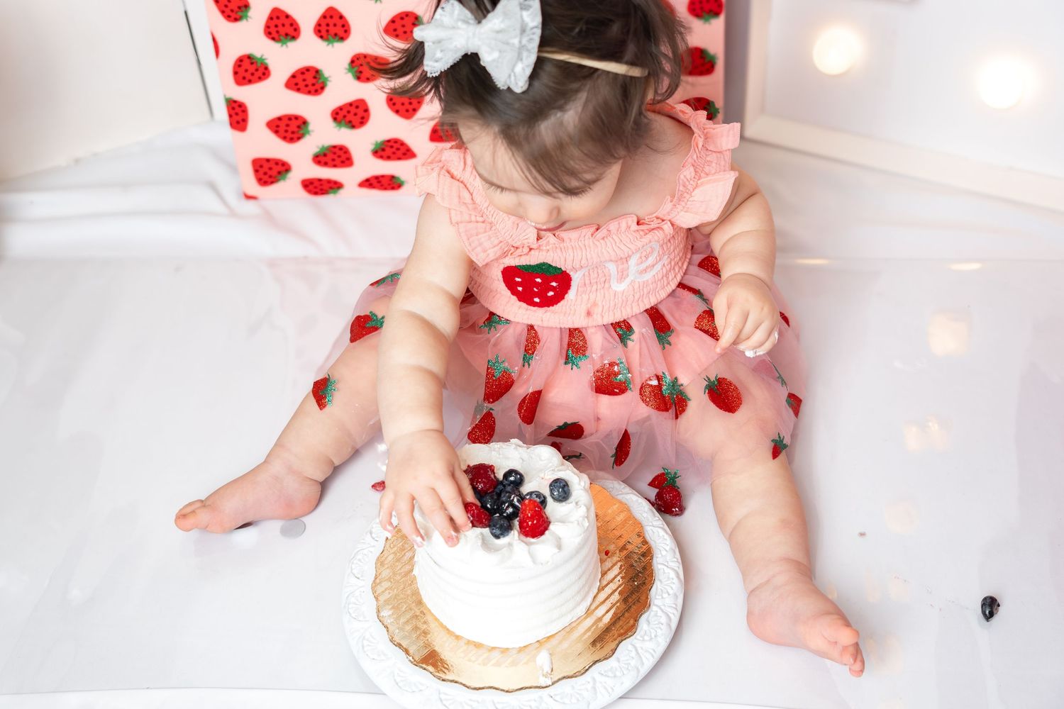 A toddler girl in a strawberry dress reaches into a white cake topped with fresh berries during a first birthday smash cake session.