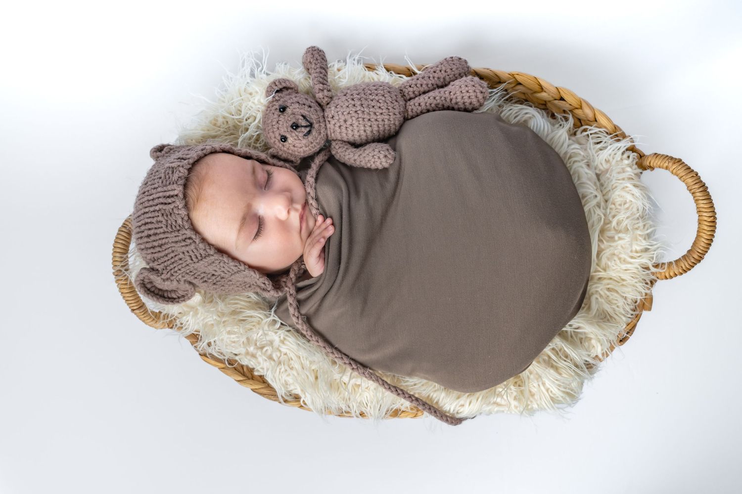 Sleeping newborn baby wrapped in brown swaddle with knit bear hat and toy in wicker basket.