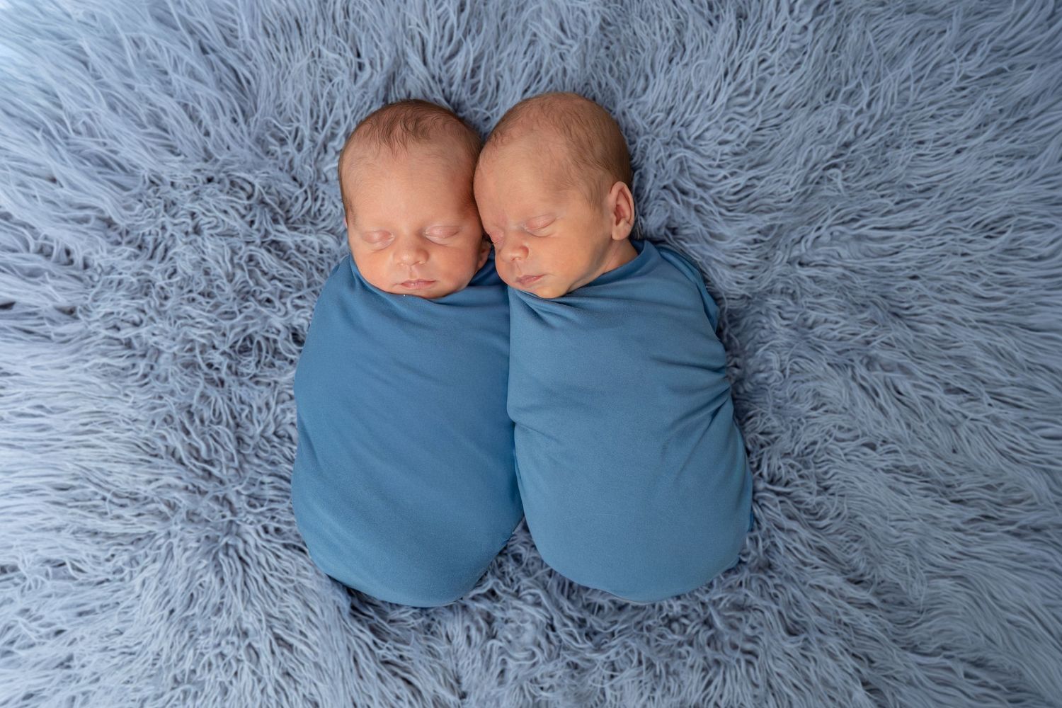 Two newborn twins swaddled in blue wraps sleeping peacefully on a soft blue fluffy blanket.
