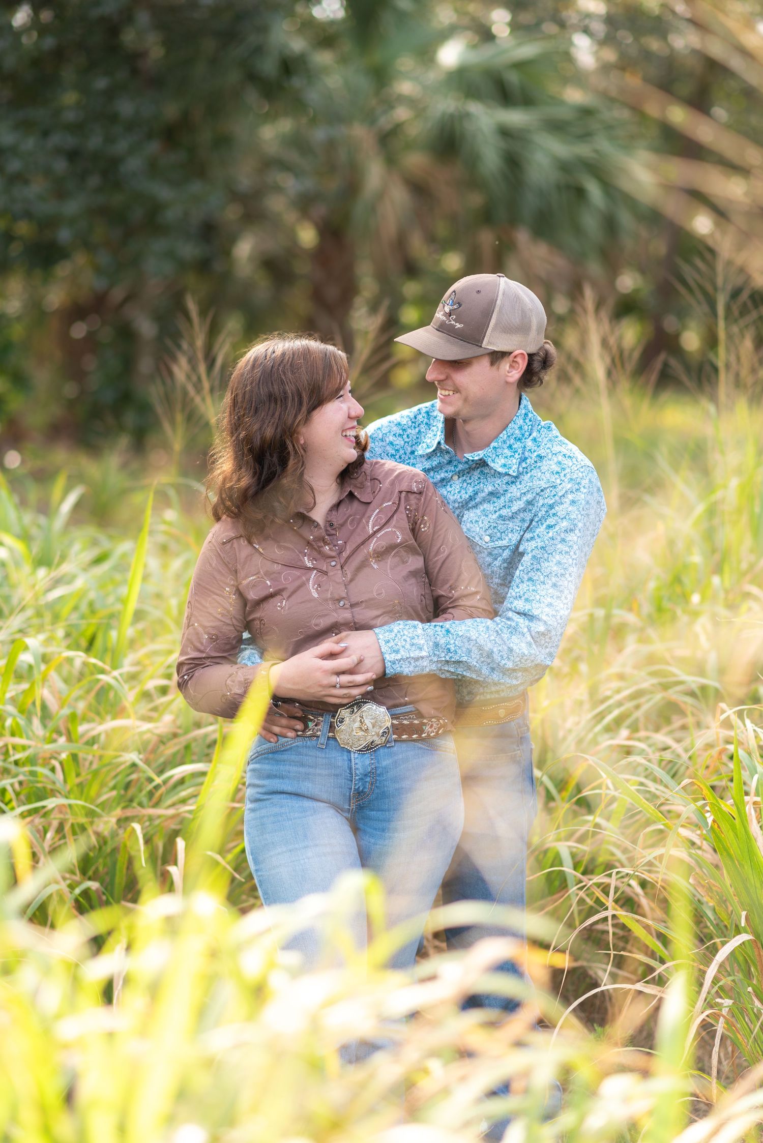Couple embracing and smiling in tall grass field, woman in brown western shirt and man in blue floral shirt with cap.