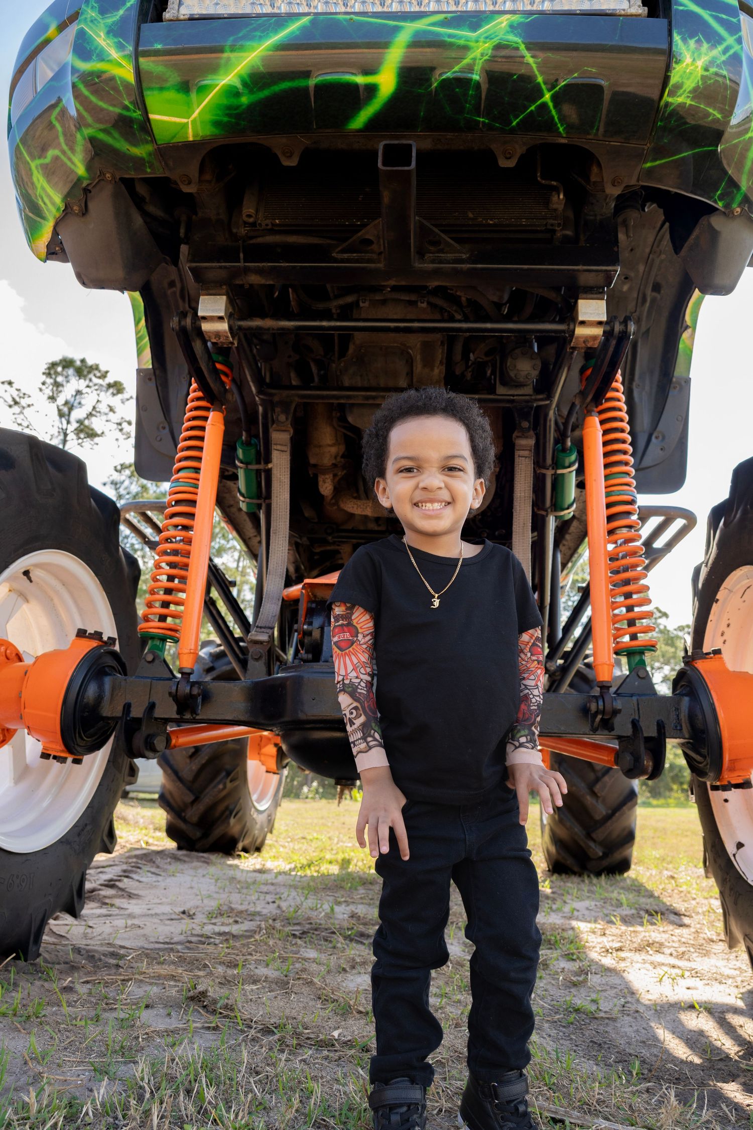 Smiling young boy in black outfit standing in front of a green monster truck with orange shocks outdoors.
