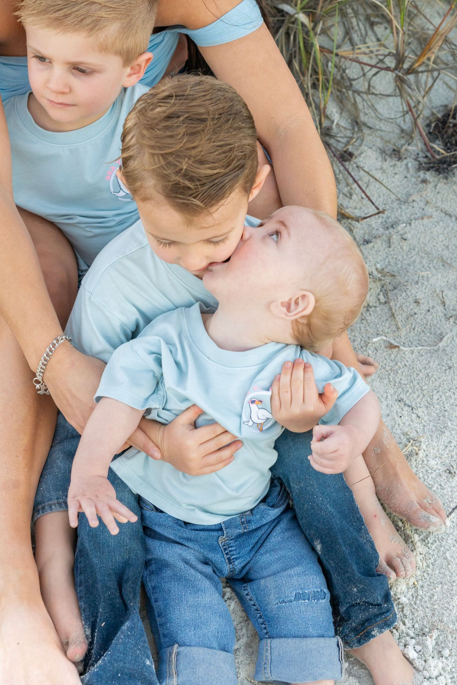 Two boys in light blue shirts sitting on sandy beach, older brother kissing baby sibling on the cheek.