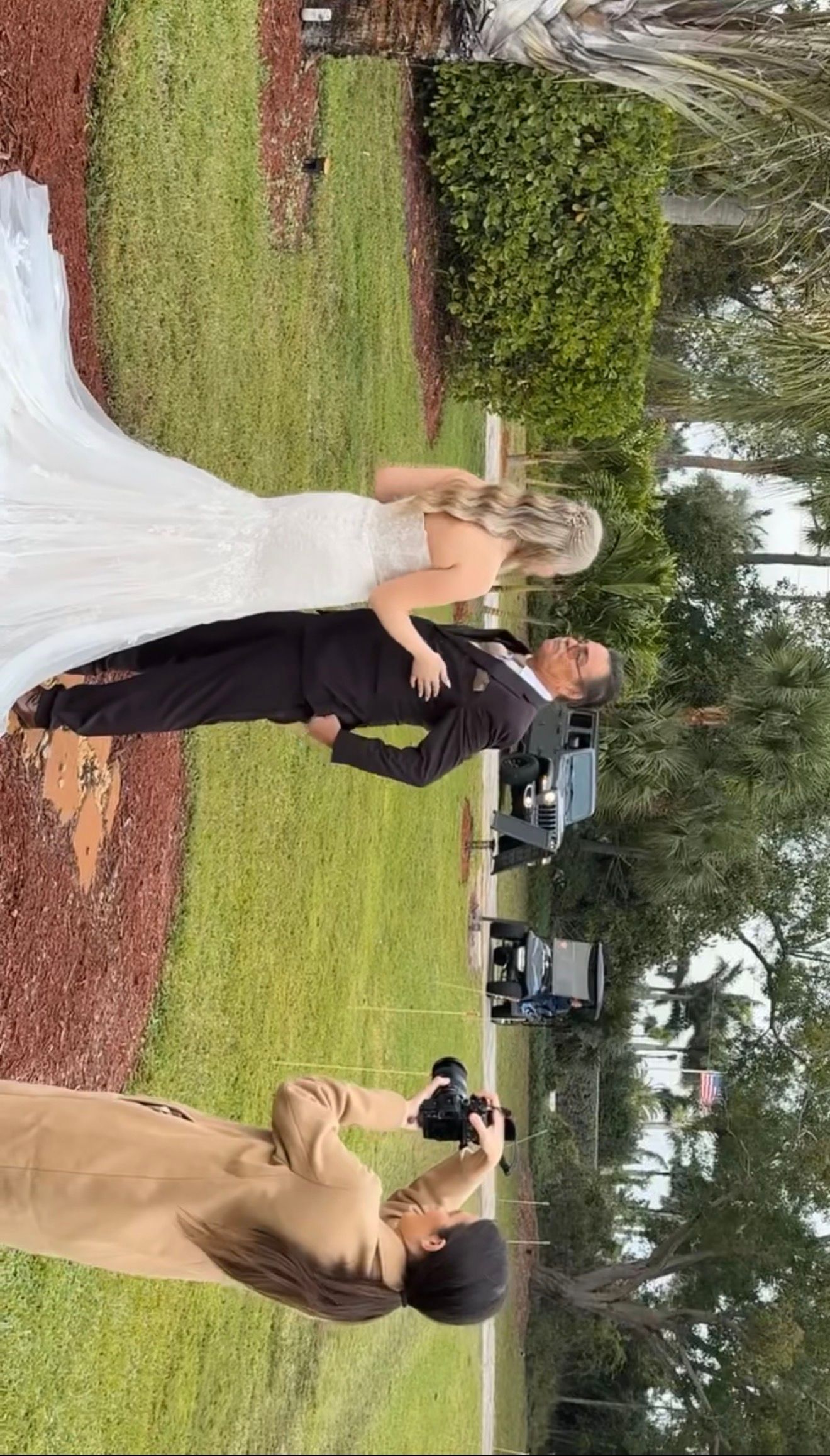 Wedding photographer crouches to capture bride and groom portrait during outdoor ceremony on grassy venue.
