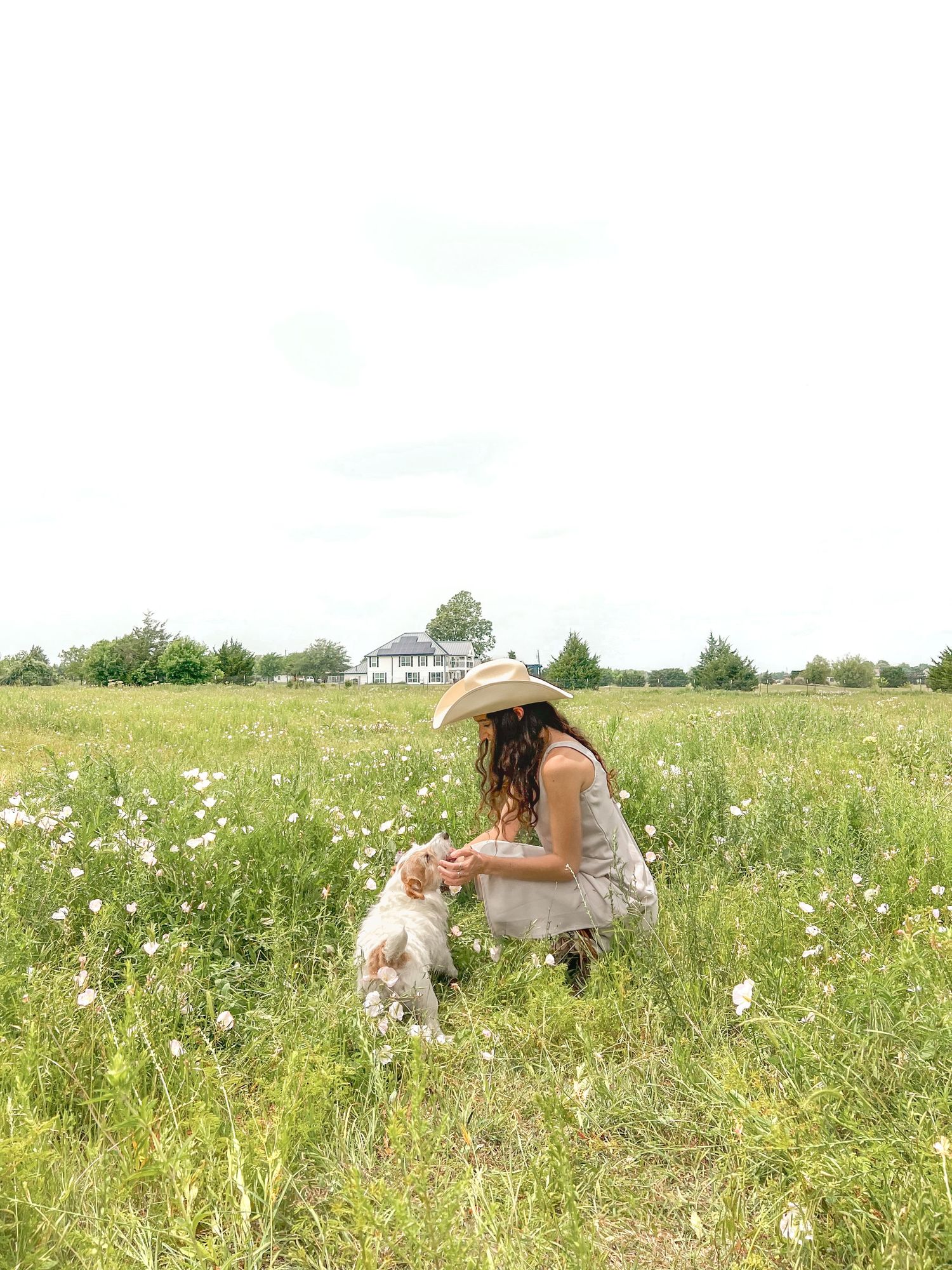 Woman in cowboy hat crouching in wildflower meadow with small dog on sunny day.