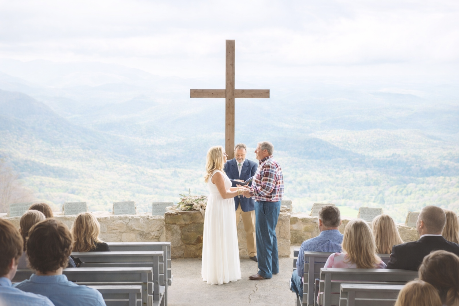Outdoor mountain wedding ceremony with couple exchanging vows before large cross with scenic valley views.