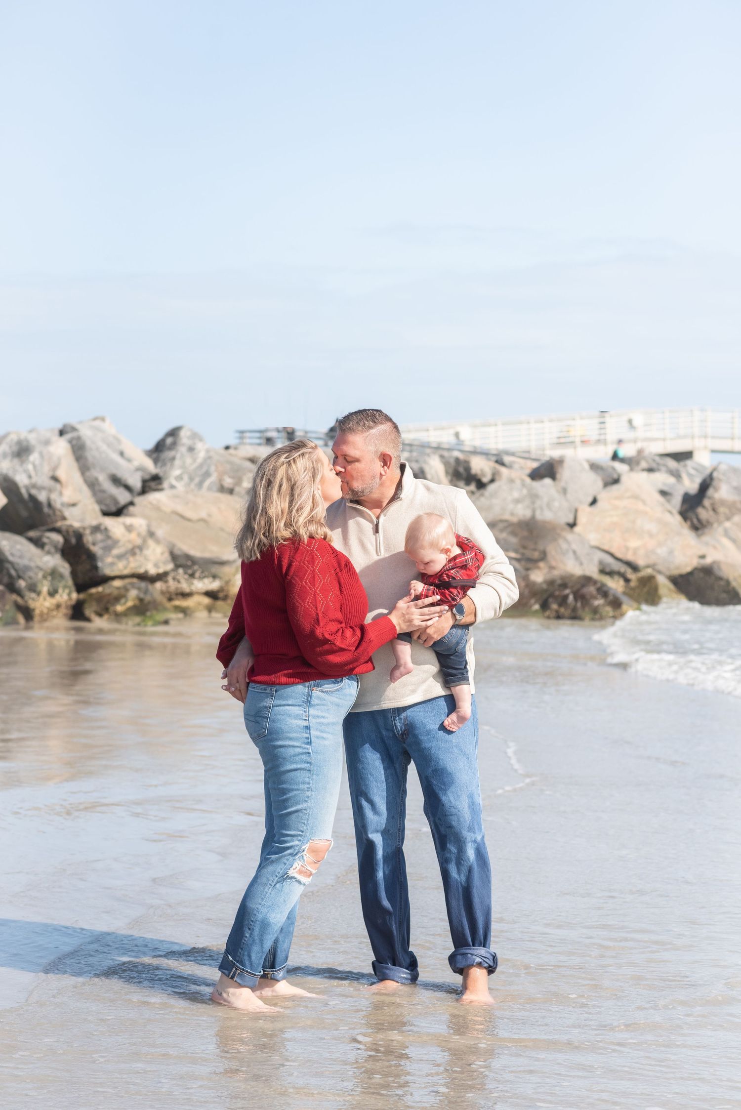 A couple sharing a kiss on the beach while holding their baby, wearing red and beige sweaters with jeans.