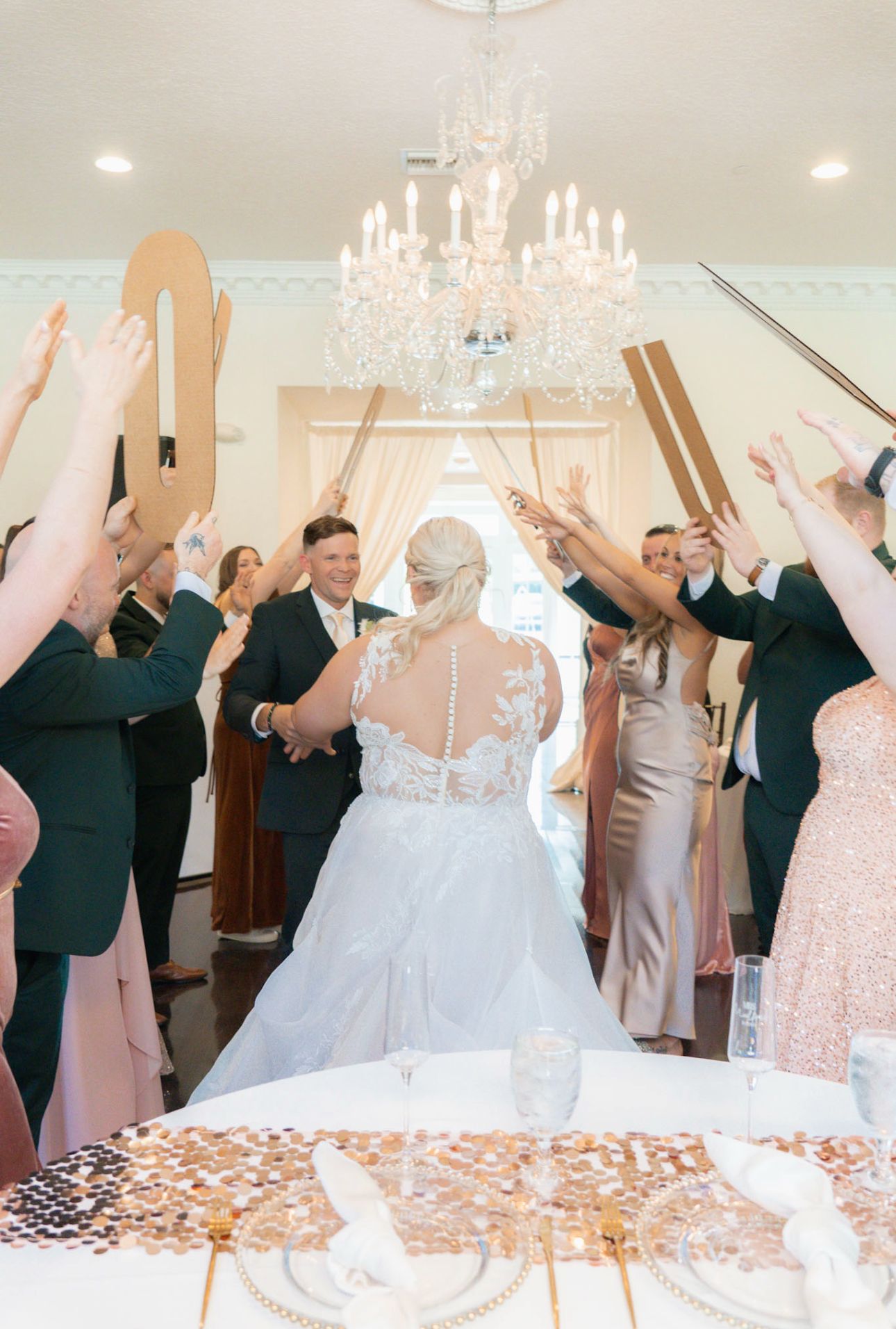 Bride and groom walk through a paddle arch under a crystal chandelier during elegant indoor wedding reception celebration.