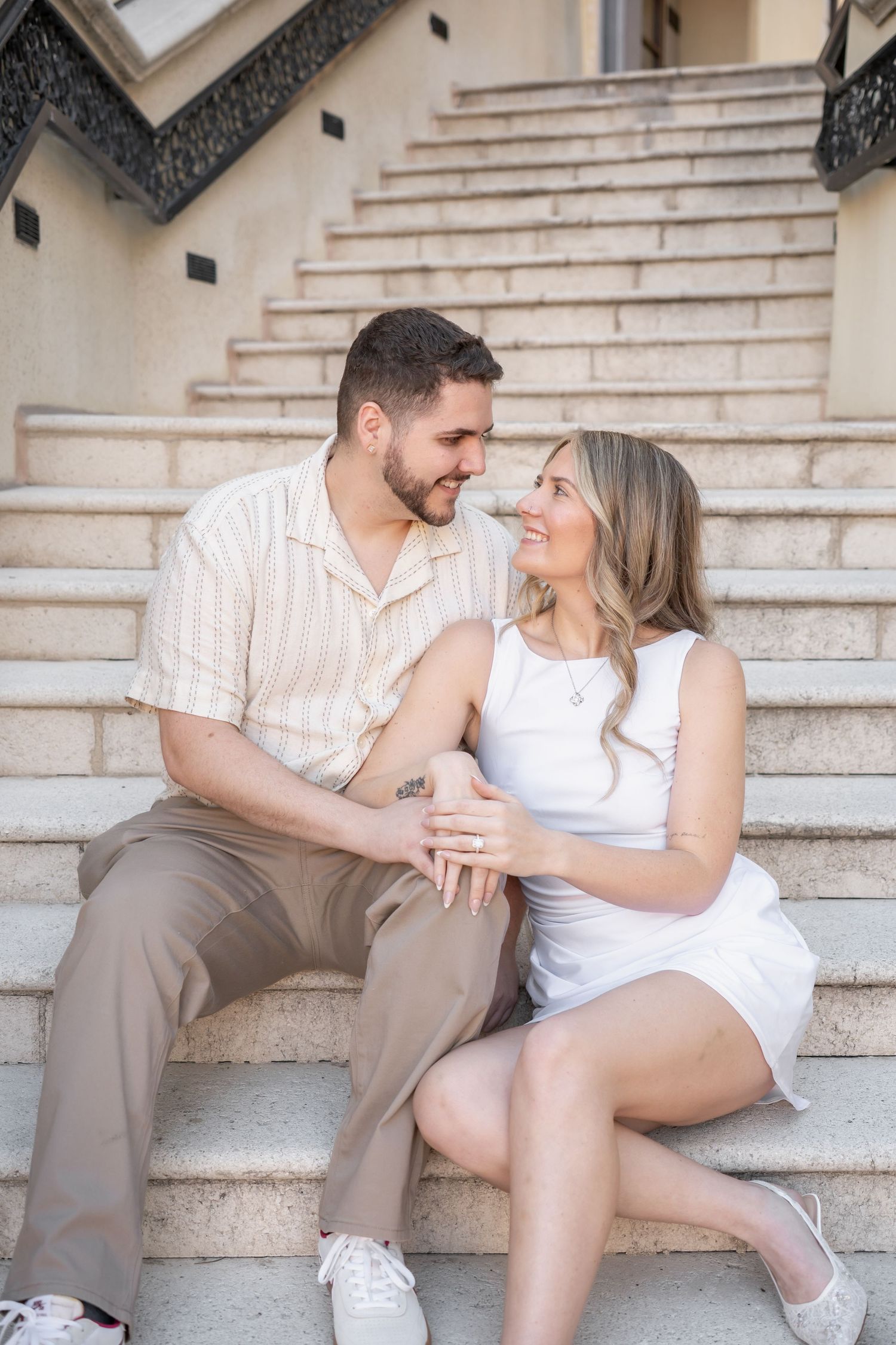 Couple sitting on stone steps gazing at each other lovingly, woman wearing white dress, man in striped shirt.