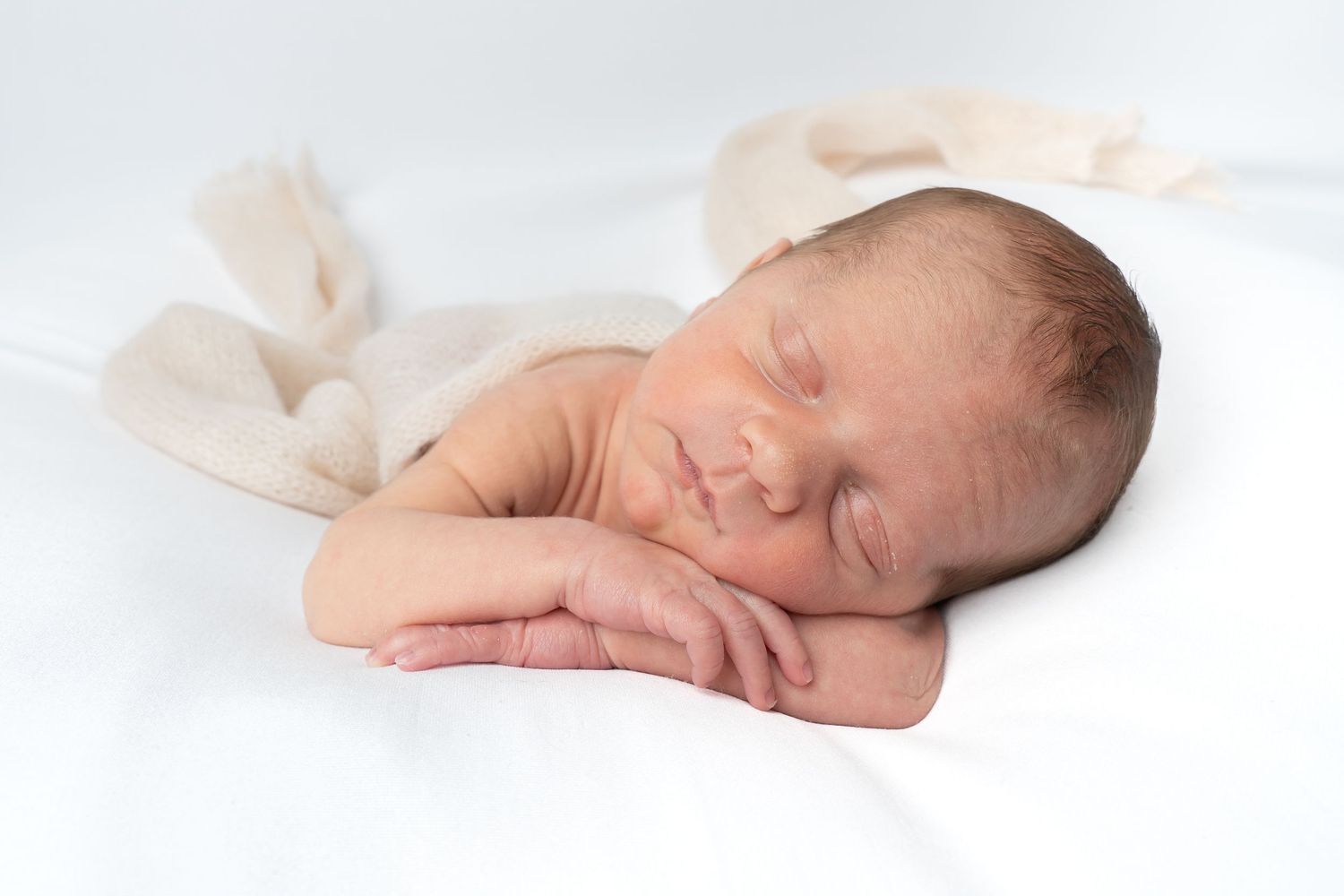 Peaceful newborn baby sleeping on white blanket with hands tucked under cheek in soft natural light.