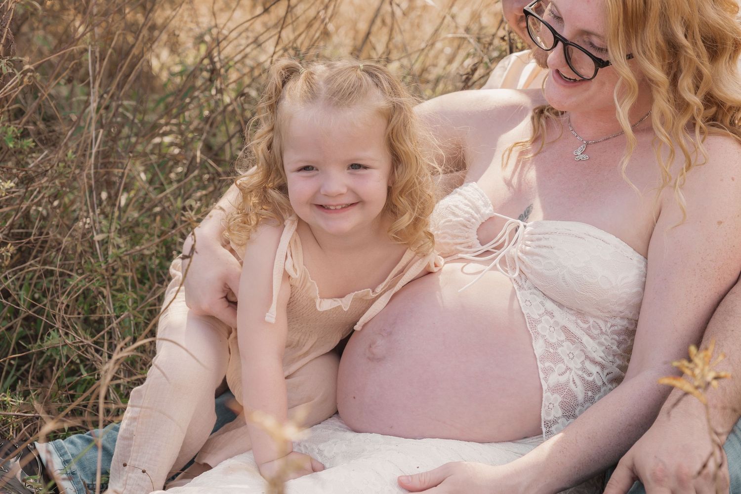 Pregnant woman sitting outdoors with smiling curly-haired toddler girl in warm golden light maternity photo.