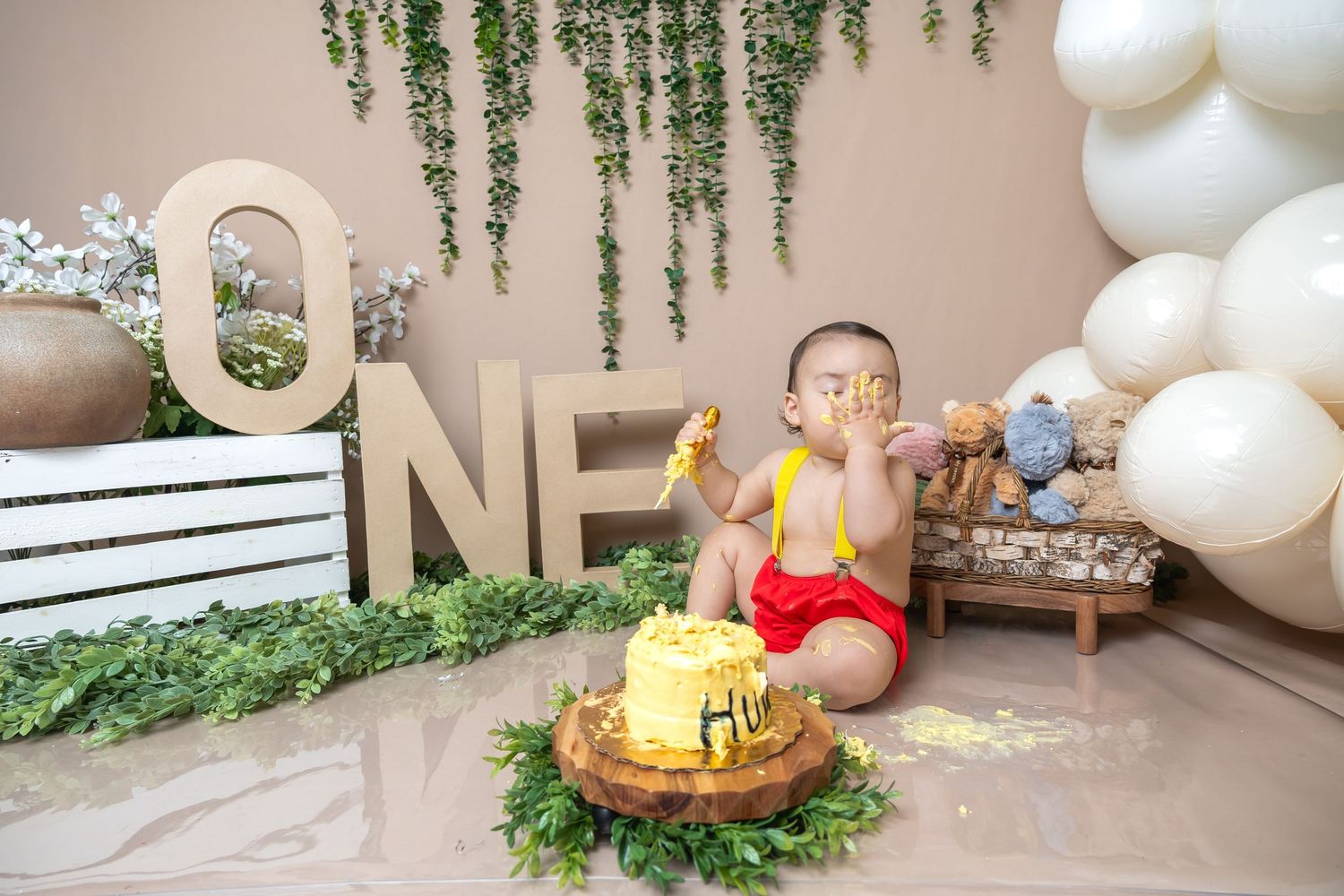 Baby boy in red shorts smashes yellow first birthday cake surrounded by ONE letters, balloons, and greenery decor.