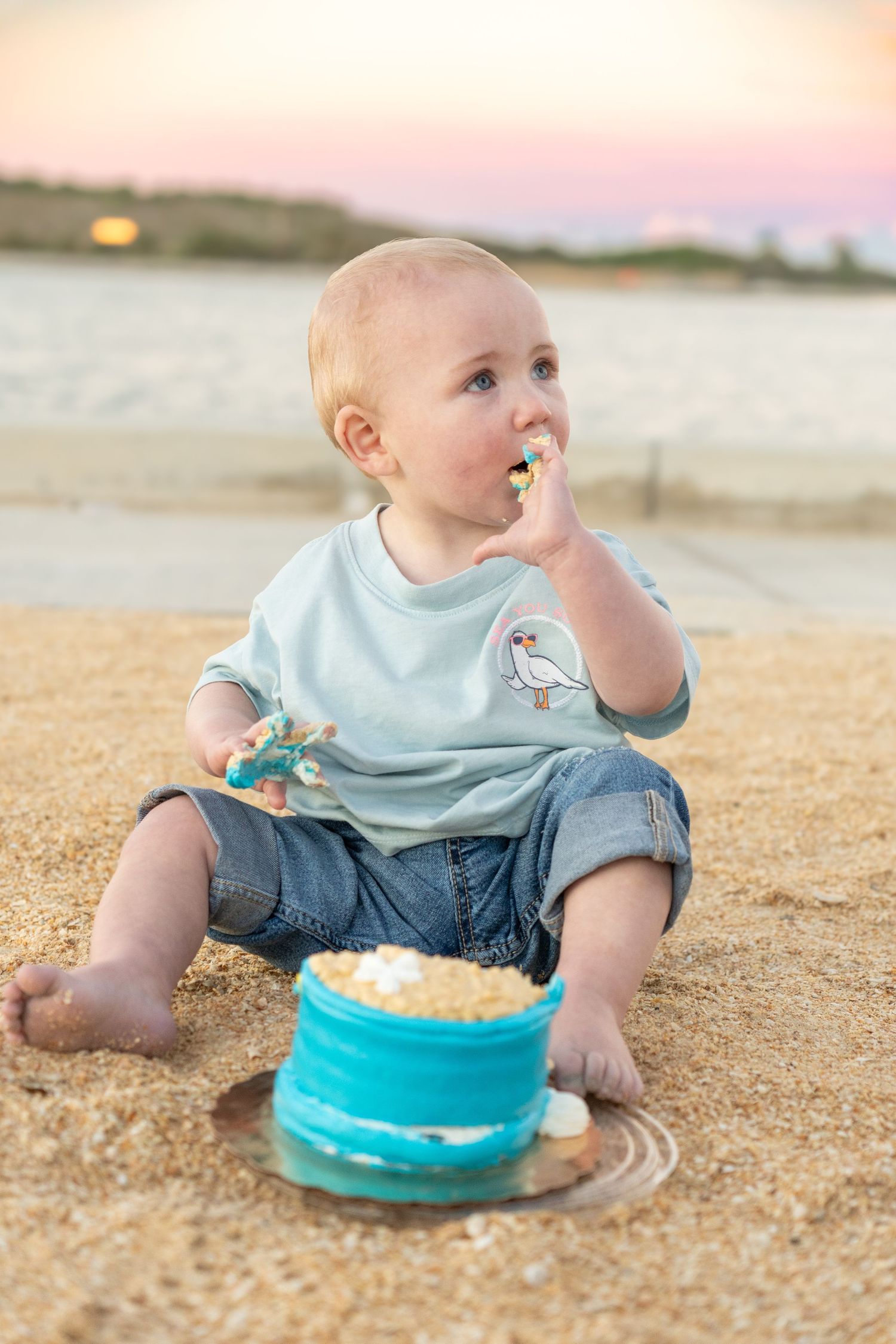 Baby boy sitting on sandy beach eating first birthday cake smash at sunset near water.